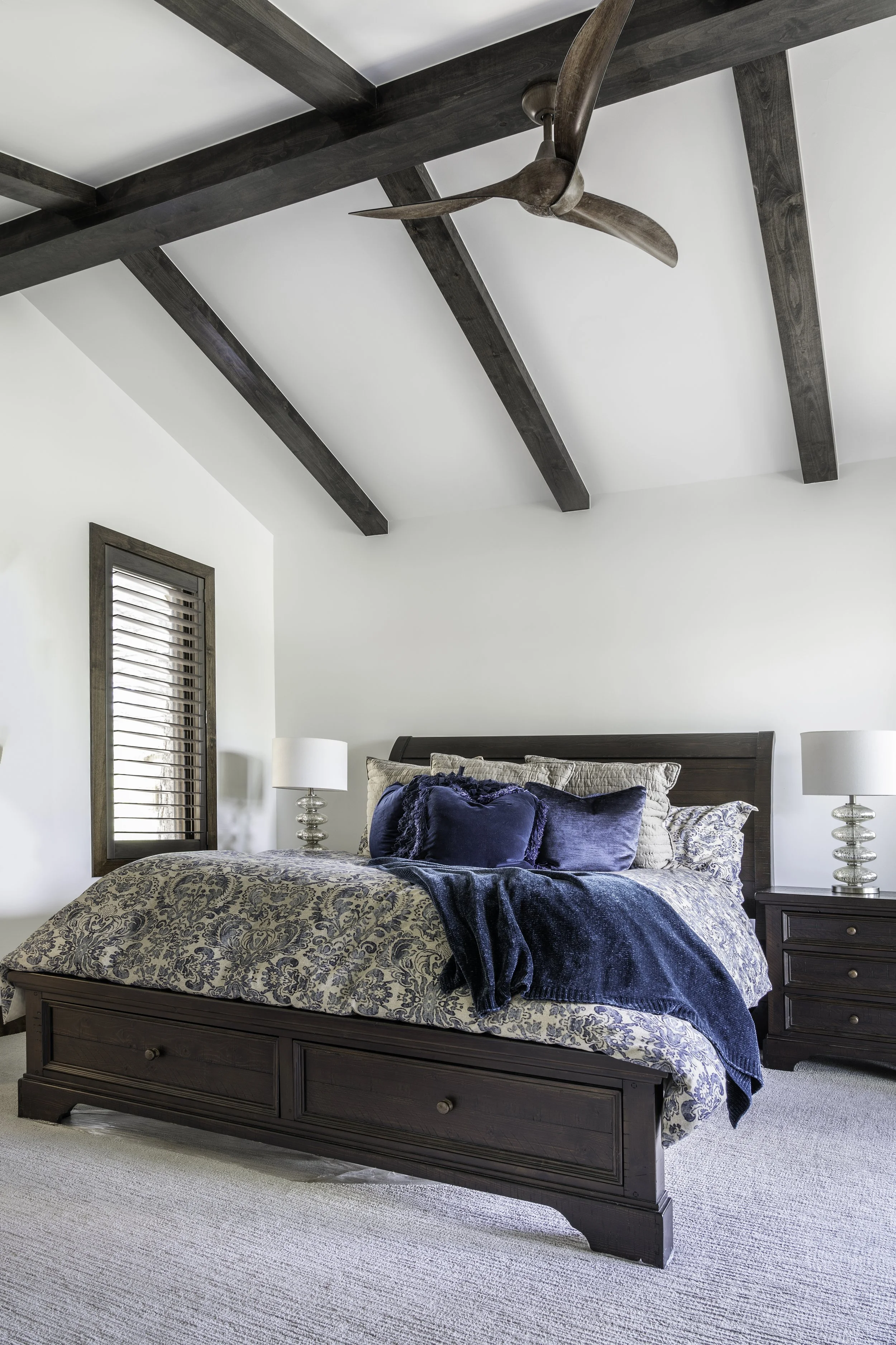 A bedroom with a dark wooden bed frame, blue and beige patterned bedding, and a dark blue throw. There are two nightstands with white lamps, and a wooden ceiling fan with three blades. The room has white walls, a small window with wooden blinds, and 