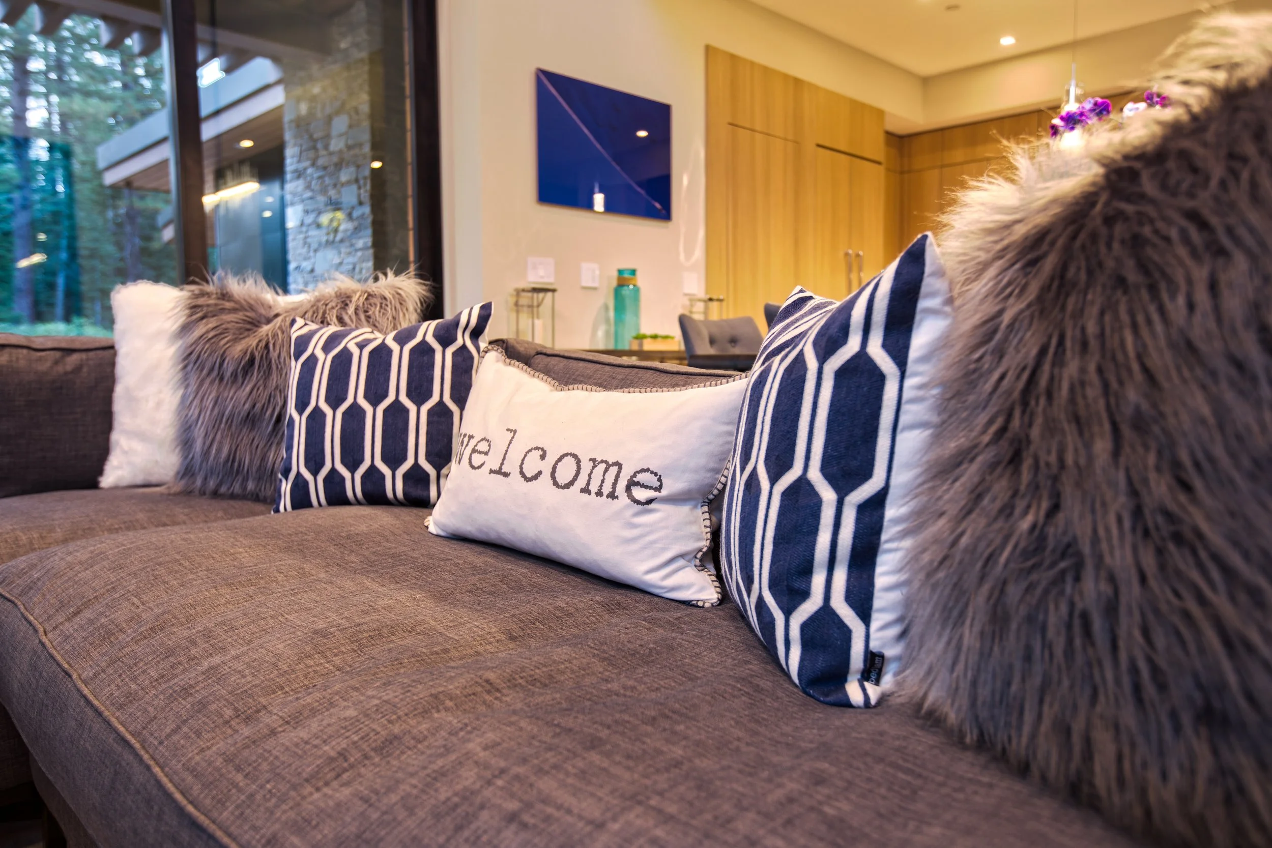 Close-up of decorative pillows on a brown fabric sofa, including a white pillow with 'welcome' embroidered on it and others with geometric and faux fur patterns, in a modern living room with a wood accent wall and a mounted flat-screen TV.