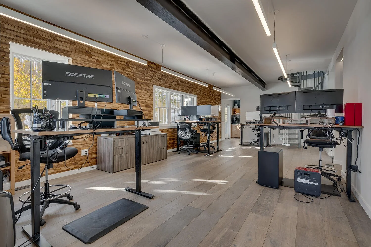 Modern office with multiple desks, ergonomic chairs, dual monitors, and natural light from windows. Wooden wall accent and sleek lighting fixtures.