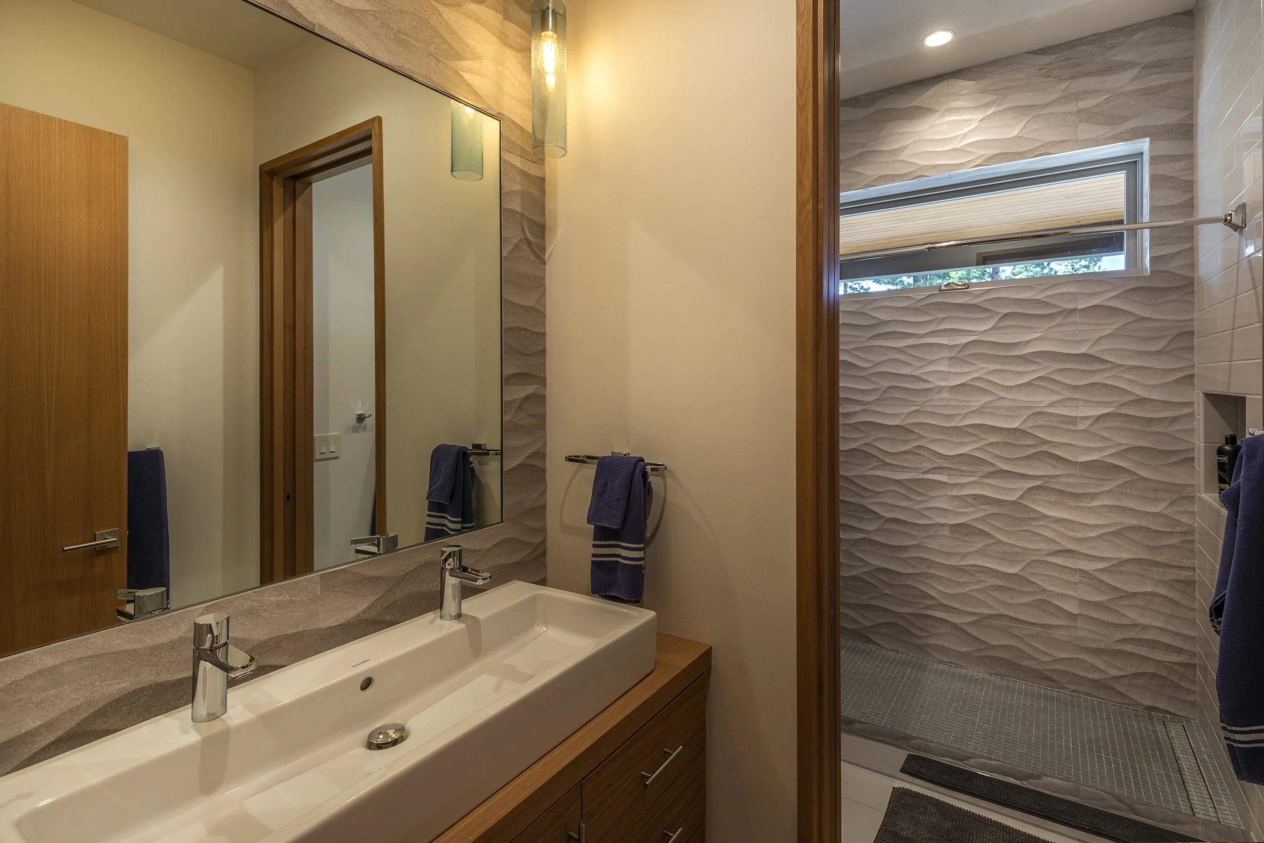 Modern bathroom with double sink vanity, large mirror, wall-mounted towel racks with navy towels, and a walk-in shower with textured tile walls and a small horizontal window.