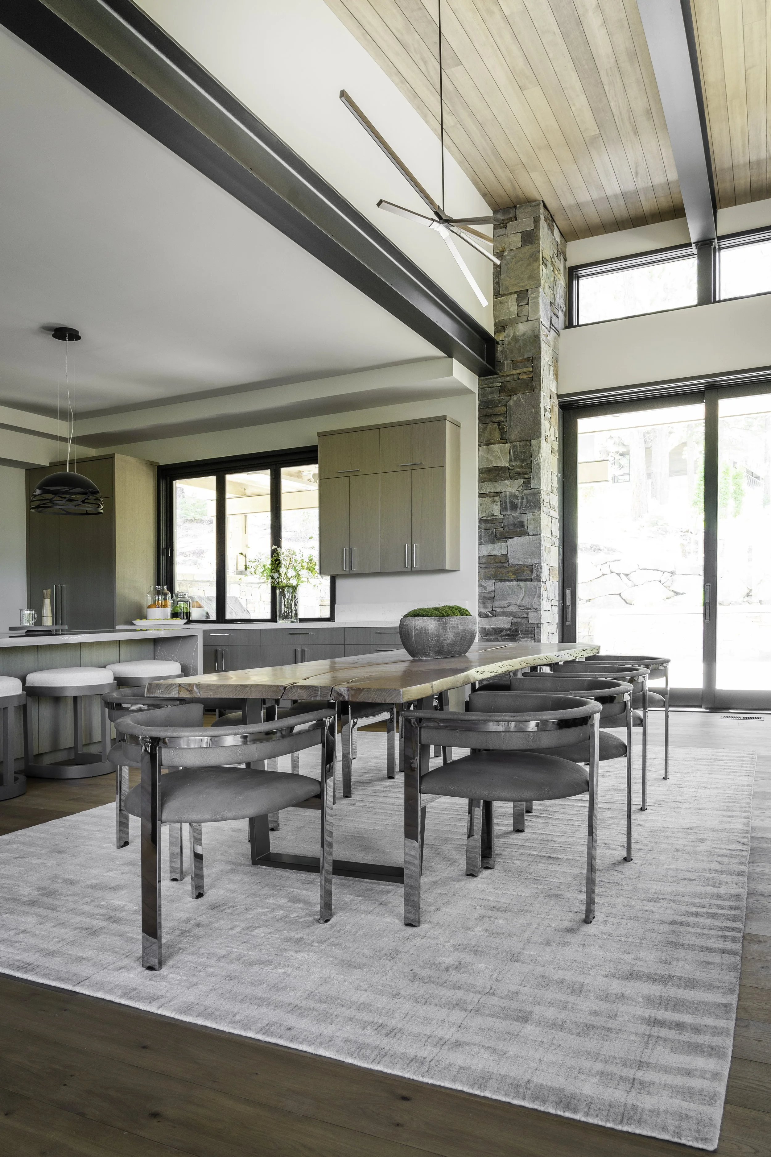 Modern dining area with a wooden table, four gray chairs, a gray and white rug, and large windows in a bright, open space.
