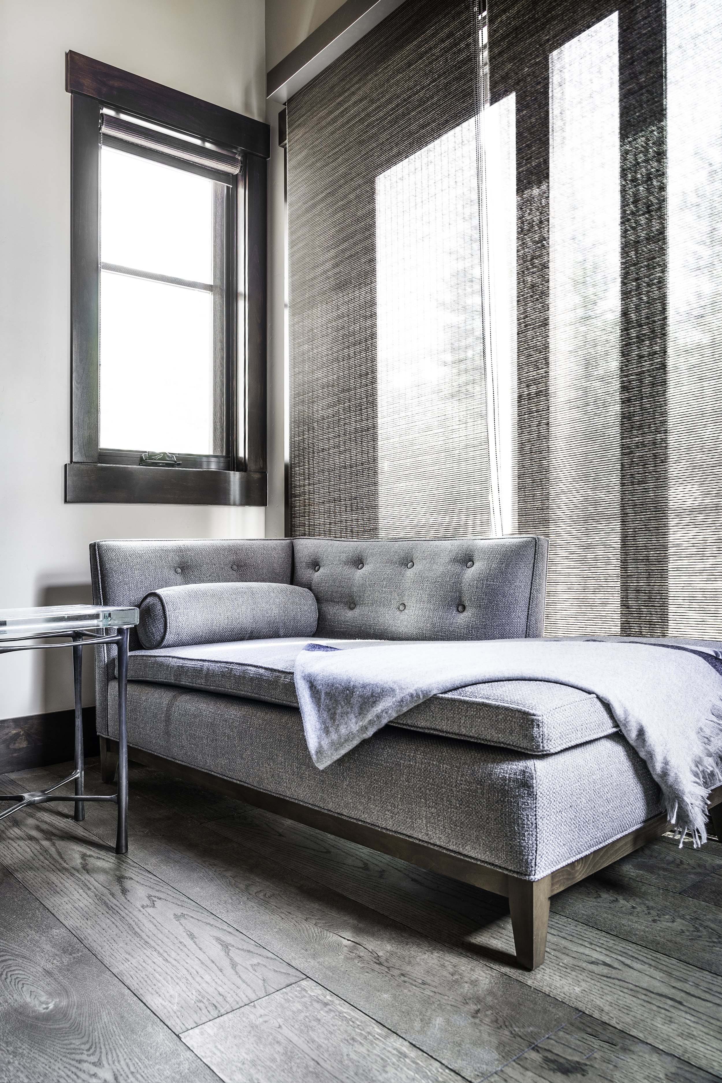 A cozy living room corner features a gray upholstered couch with a matching throw blanket and a bolster pillow. Next to the couch is a small glass side table with a chrome frame. Behind the couch, large windows with black trim let in natural light, w