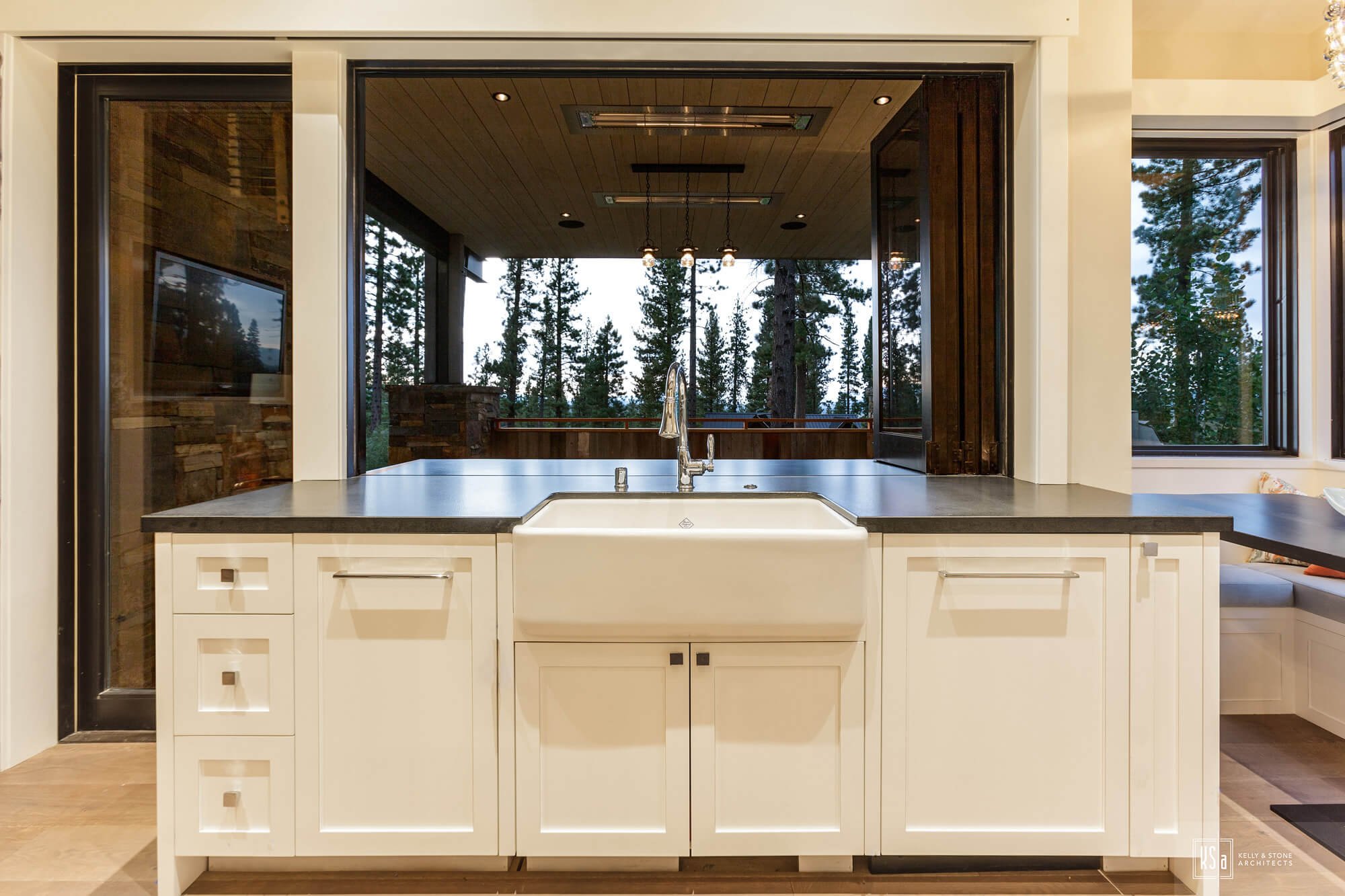 Kitchen sink with white cabinetry and a large window above, overlooking a forested outdoor space.