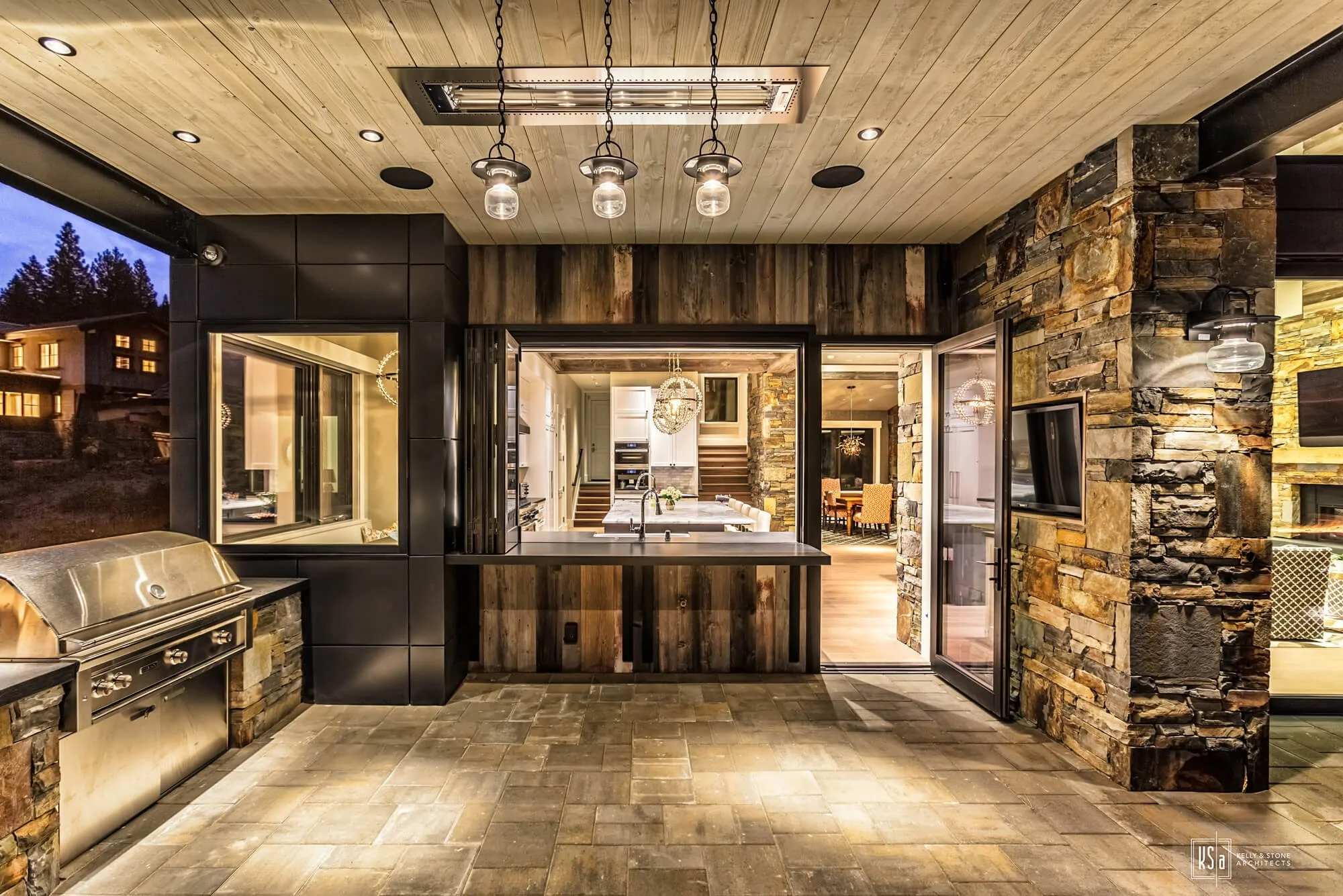 Exterior patio area with stone, wood, and black paneling, featuring a built-in stainless steel grill, pendant lights on the ceiling, and an open sliding door leading to a modern kitchen and dining area.