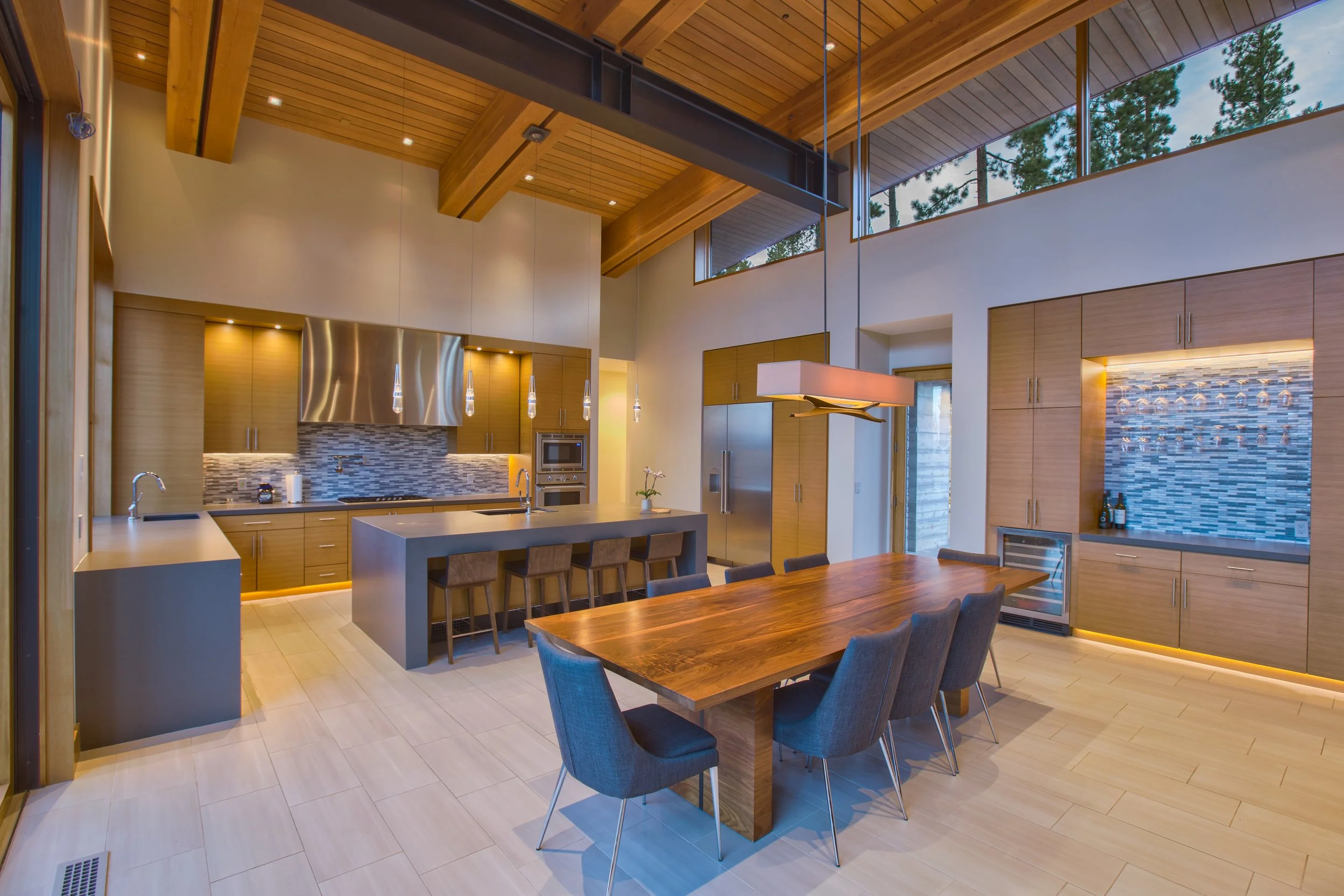 Modern kitchen and dining area with wooden ceiling and ceiling-high windows showing trees outside, featuring a kitchen island with bar stools, stainless steel appliances, and a wooden dining table with chairs.