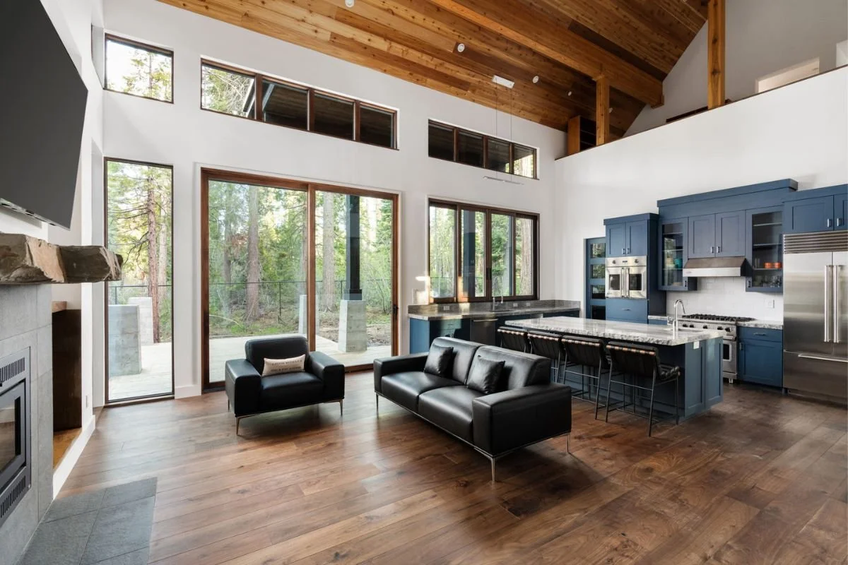 Interior of a modern open-plan living room and kitchen with large windows, wooden ceiling, black leather sofas, and blue kitchen cabinets.