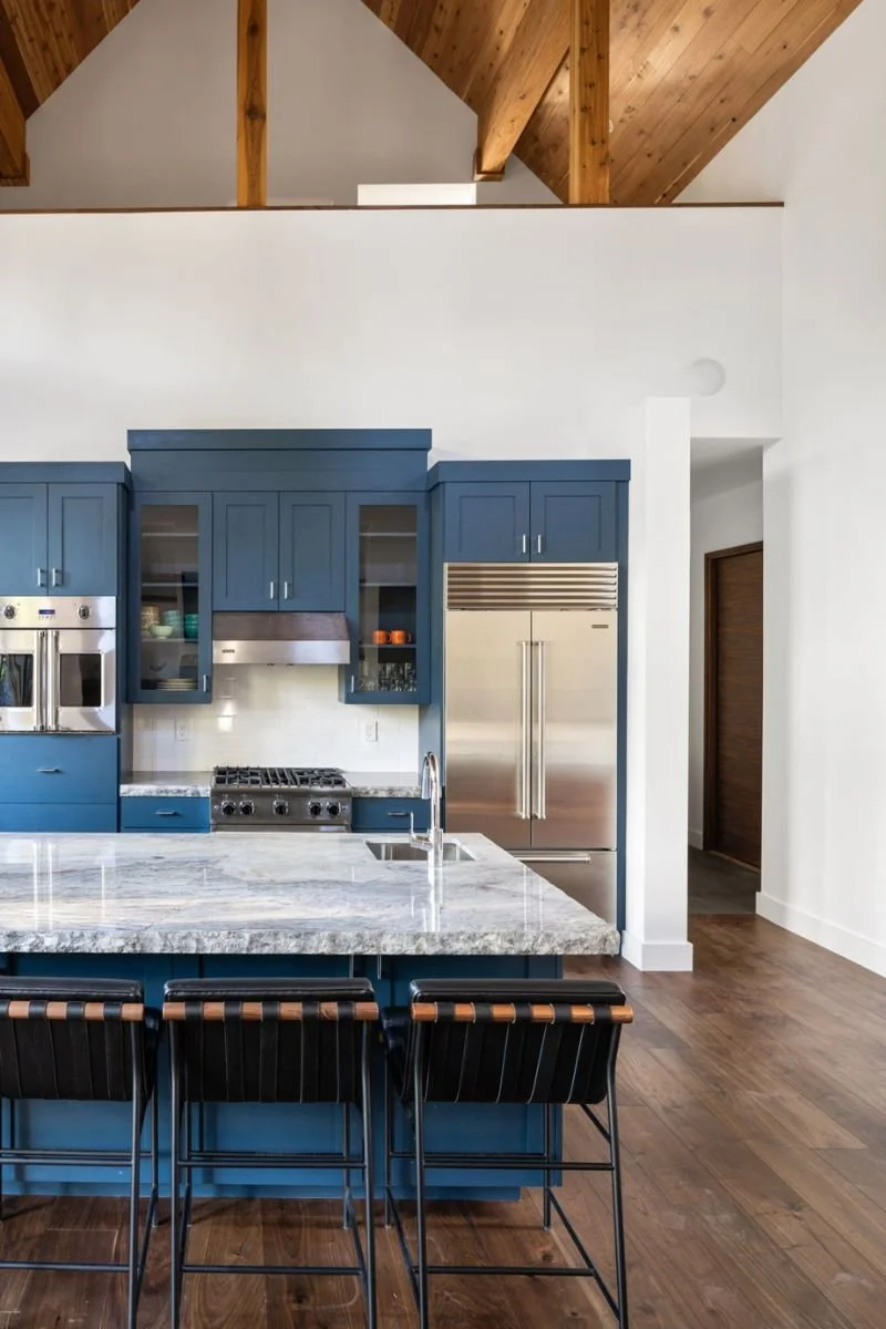 Modern kitchen with blue cabinetry, marble countertop island, and stainless steel appliances, including a large refrigerator and oven, with a high wood ceiling and hardwood floors.