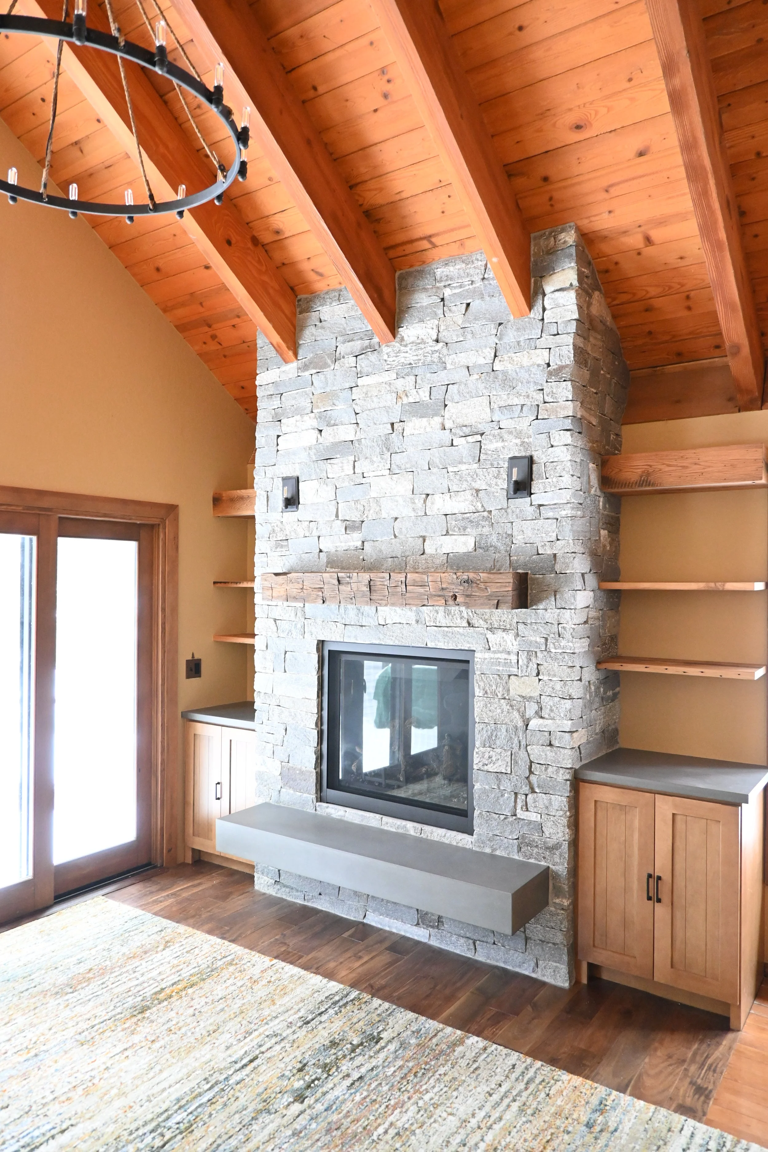 Living room corner with a stone fireplace, wooden shelves, and a wooden vaulted ceiling.