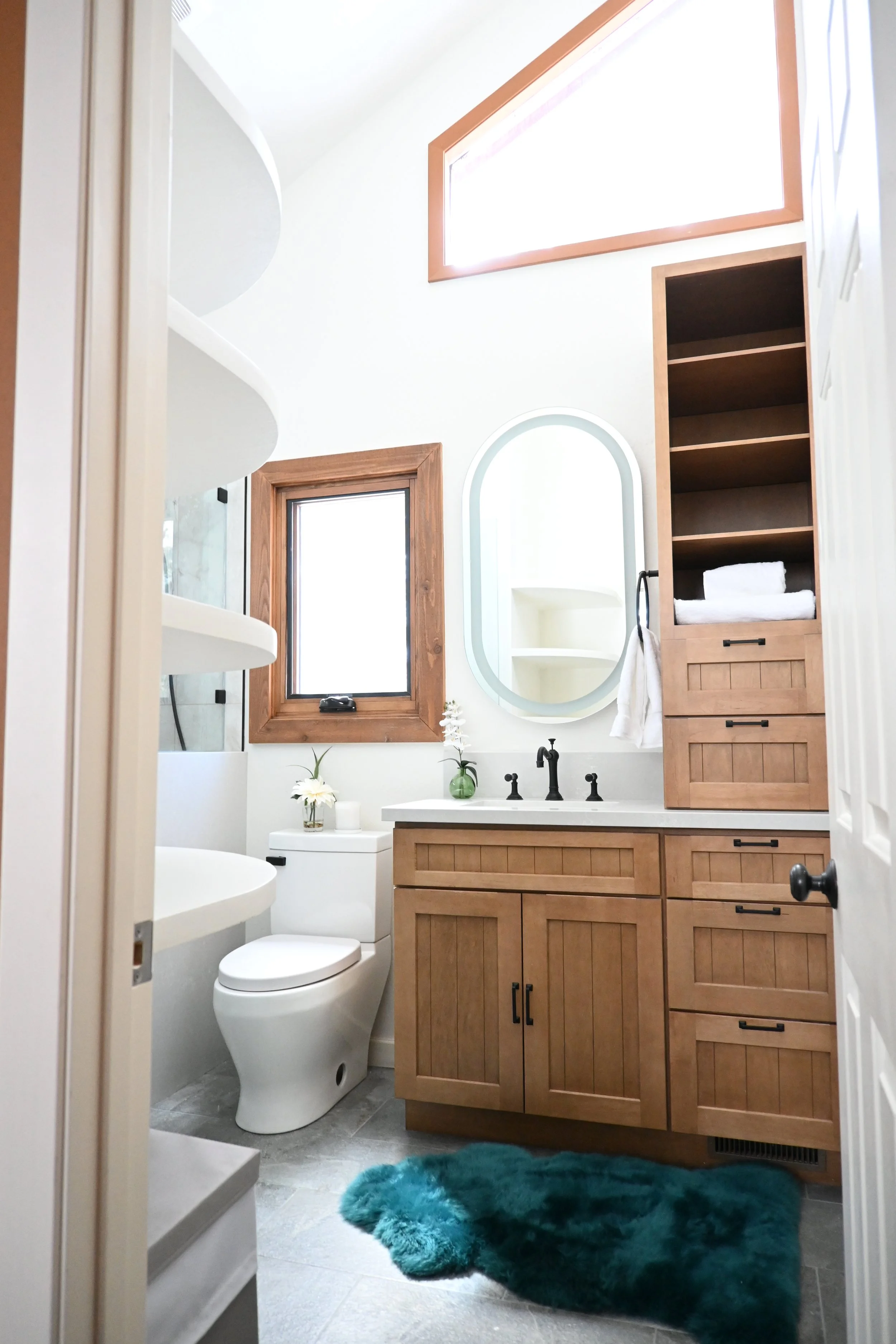 Bathroom with wood cabinets, a white toilet, and a teal rug. There are two windows, one above and one beside the toilet, and a mirror above the sink. The bathroom has light-colored walls and floors.