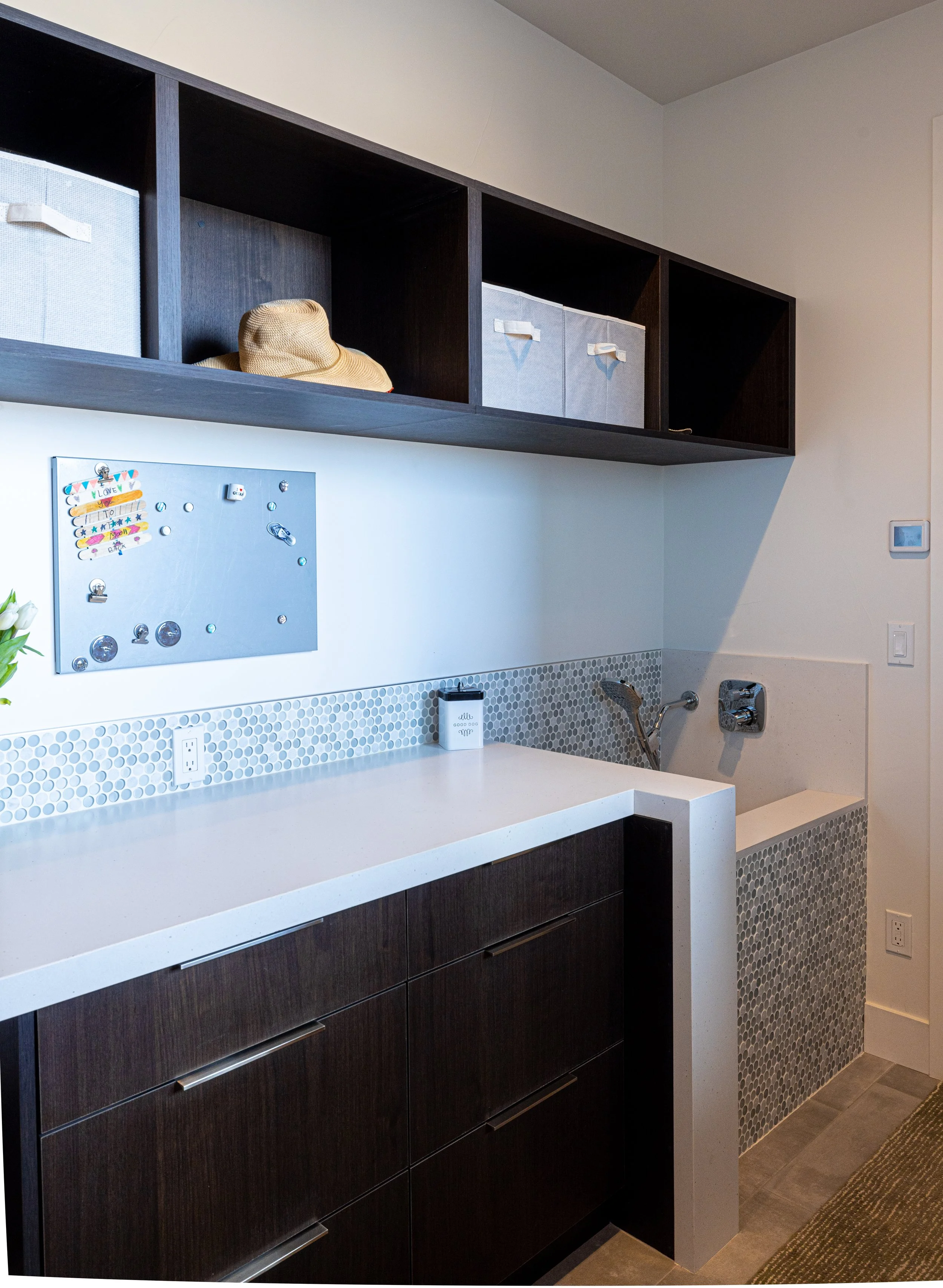 A laundry or utility room with dark wood cabinets, a white countertop, a pegboard with magnets and notes, a small decorative box, a woven hat, and a laundry sink with a metal faucet. There are storage boxes on a hi shelf and a small green plant on th