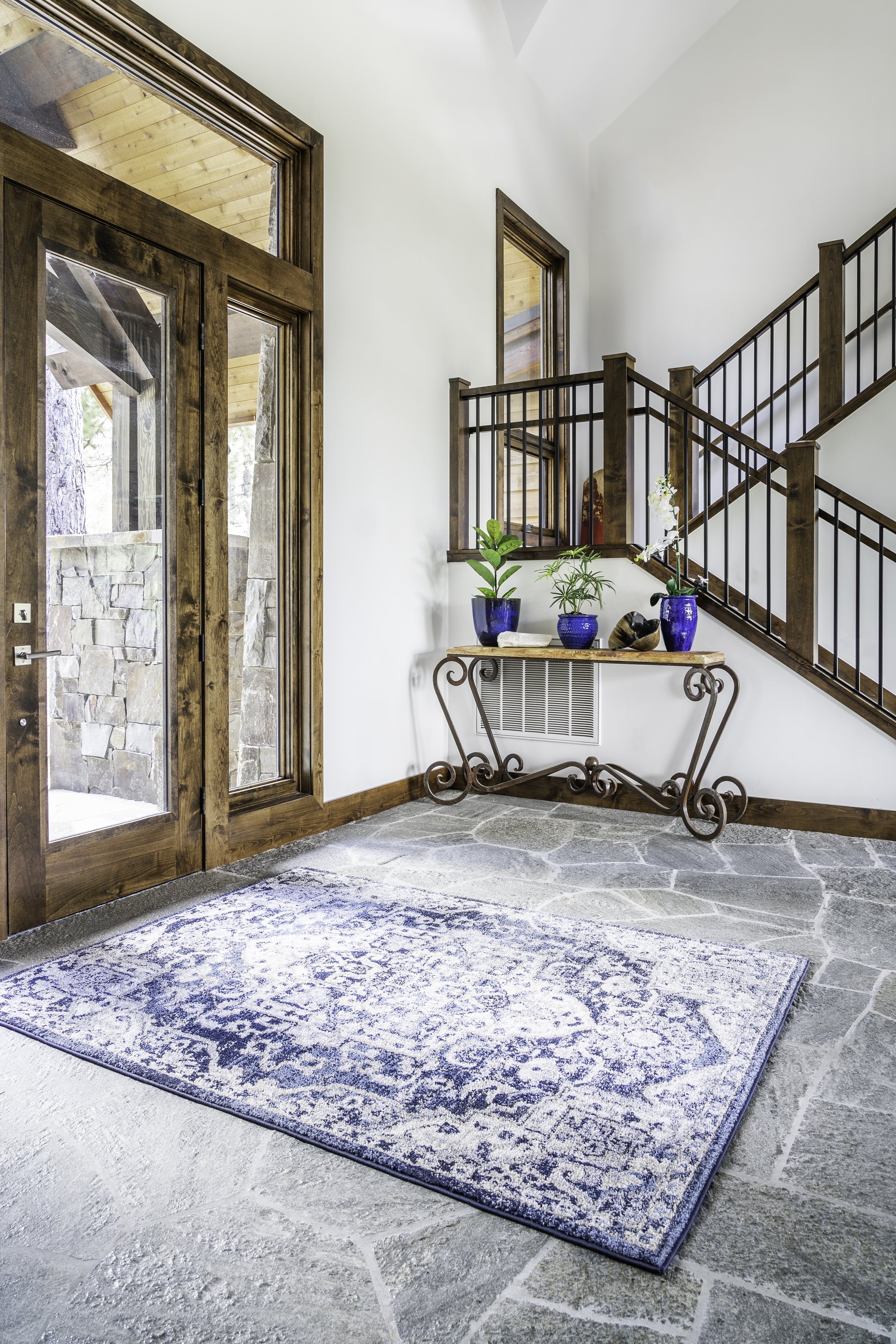Entryway of a house with a stone floor, a blue patterned rug, a wooden and iron side table with potted plants, and a staircase with wooden handrails and black balusters.