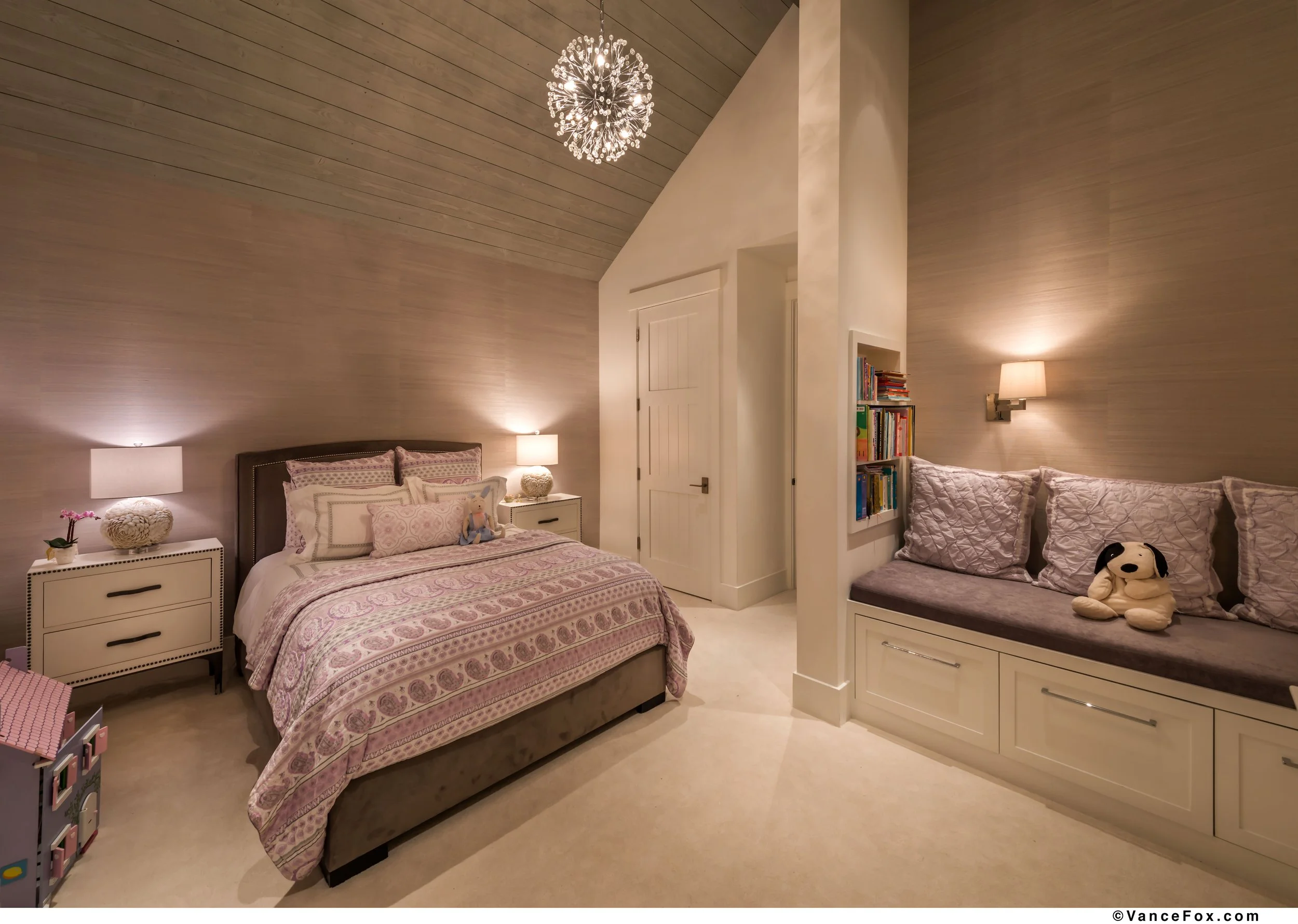 A cozy bedroom with a large bed covered in pink patterned bedding and pillows, flanked by two nightstands with lamps. There is a built-in window seat with cushions and a stuffed dog, and a bookshelf near the wall. The room has a wood-paneled sloped c