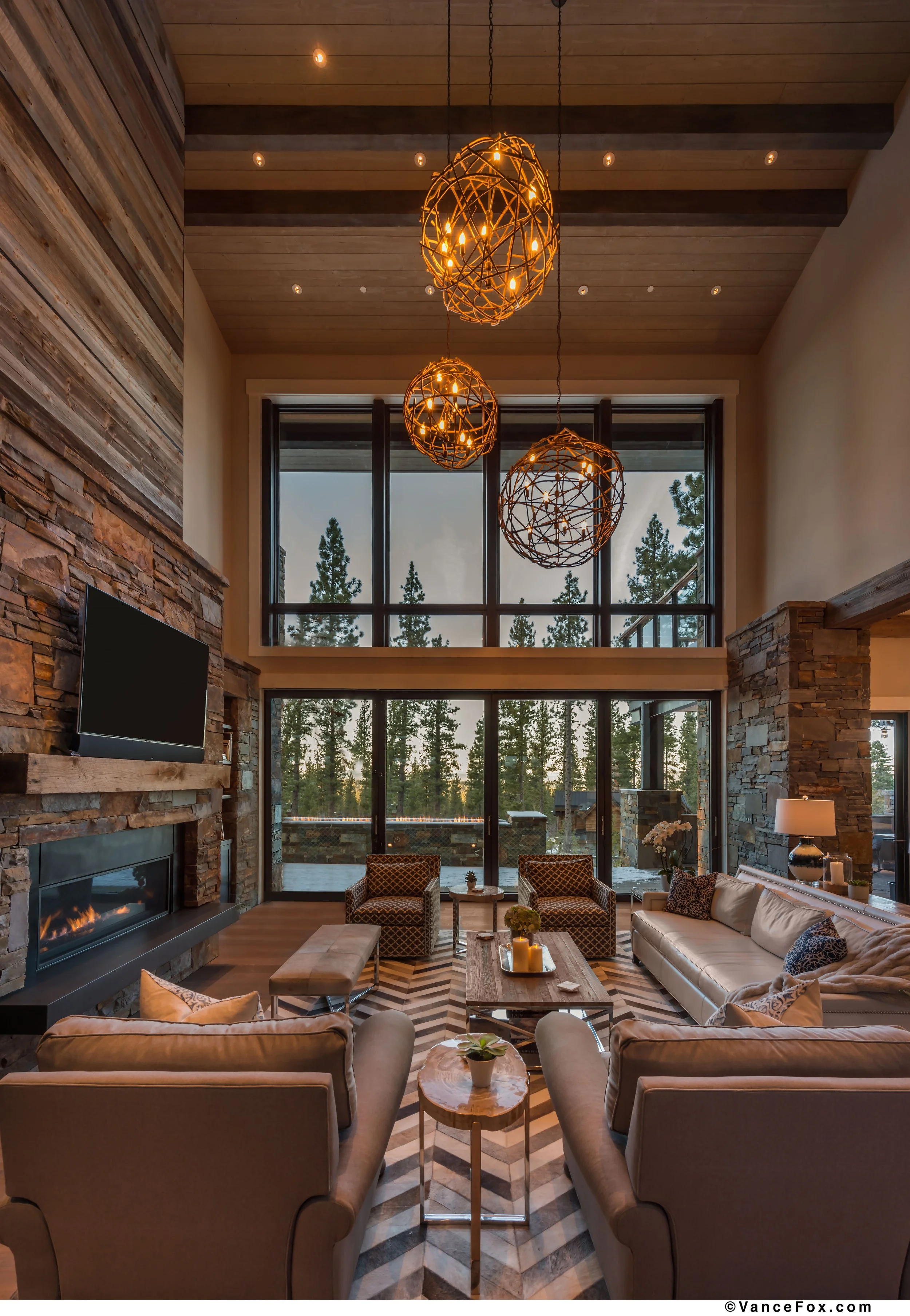 A spacious living room with high ceilings, large windows revealing pine trees outside, a stone fireplace, modern pendant lights, leather sofas, chairs, and a coffee table, creating a cozy yet modern atmosphere.