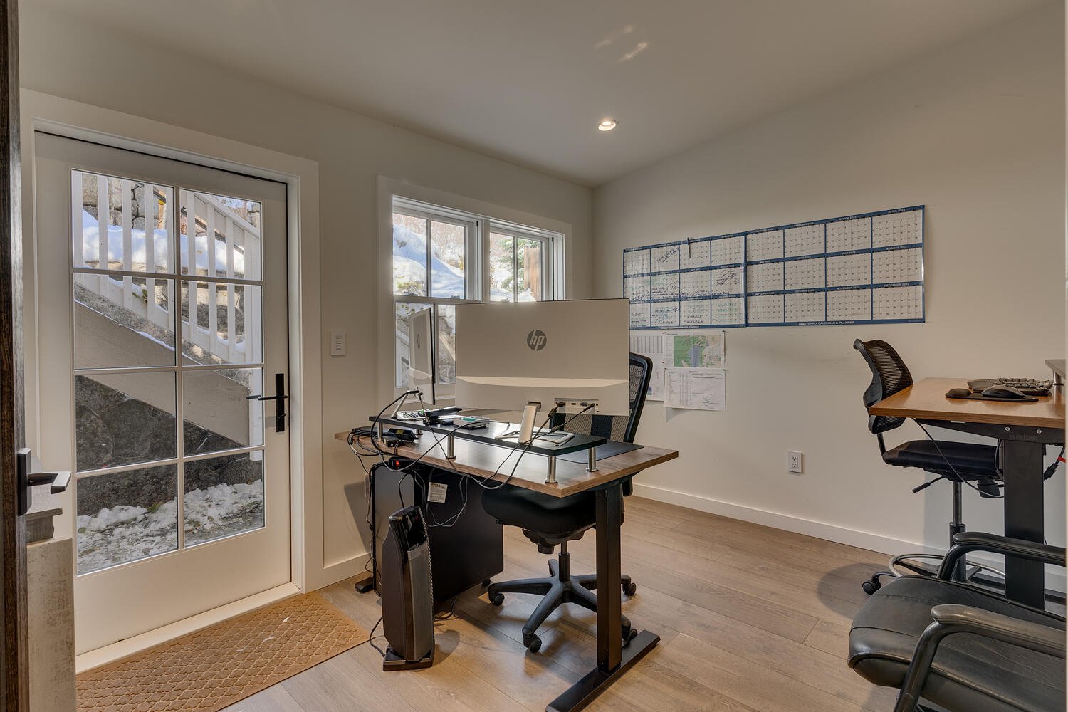 An office room with a wooden desk holding two HP monitors, a mesh office chair, and various electronic devices. Outside the glass door and window, snow-covered steps and ground are visible. A large blue and white wall calendar is hanging on the wall,