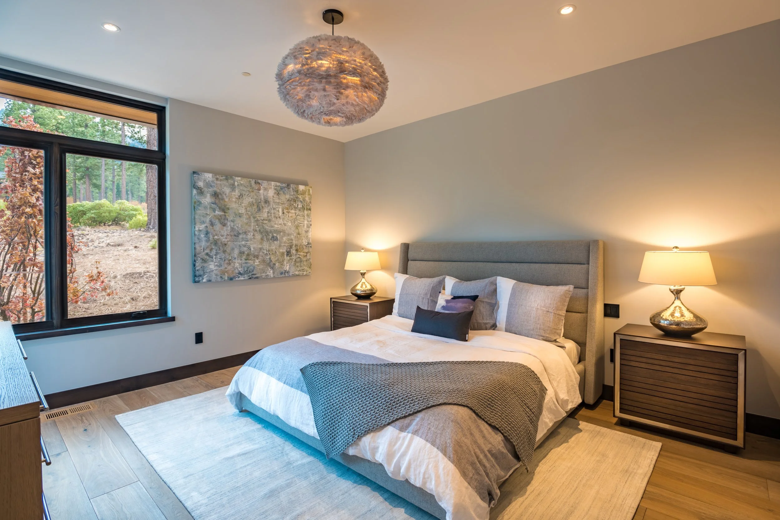 Modern bedroom with a large bed, gray upholstered headboard, covered with pillows, a blanket, and bedding. Two bedside tables with gold lamps, a window showing a natural outdoor landscape, a hanging light fixture, and artwork on the wall.