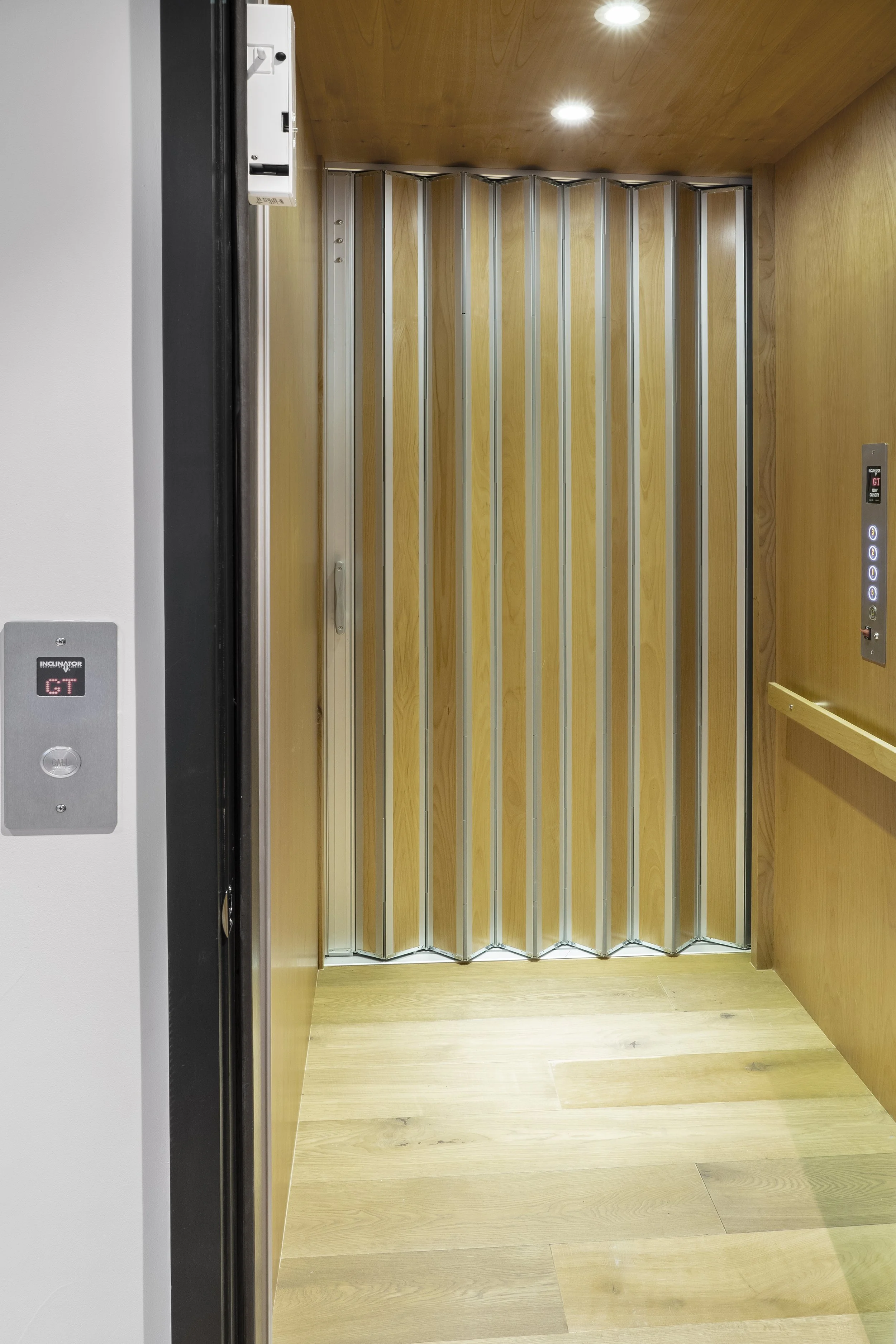 Inside an elevator with wooden walls and floor, metal folding accordion-style doors, and control panels on the right side.