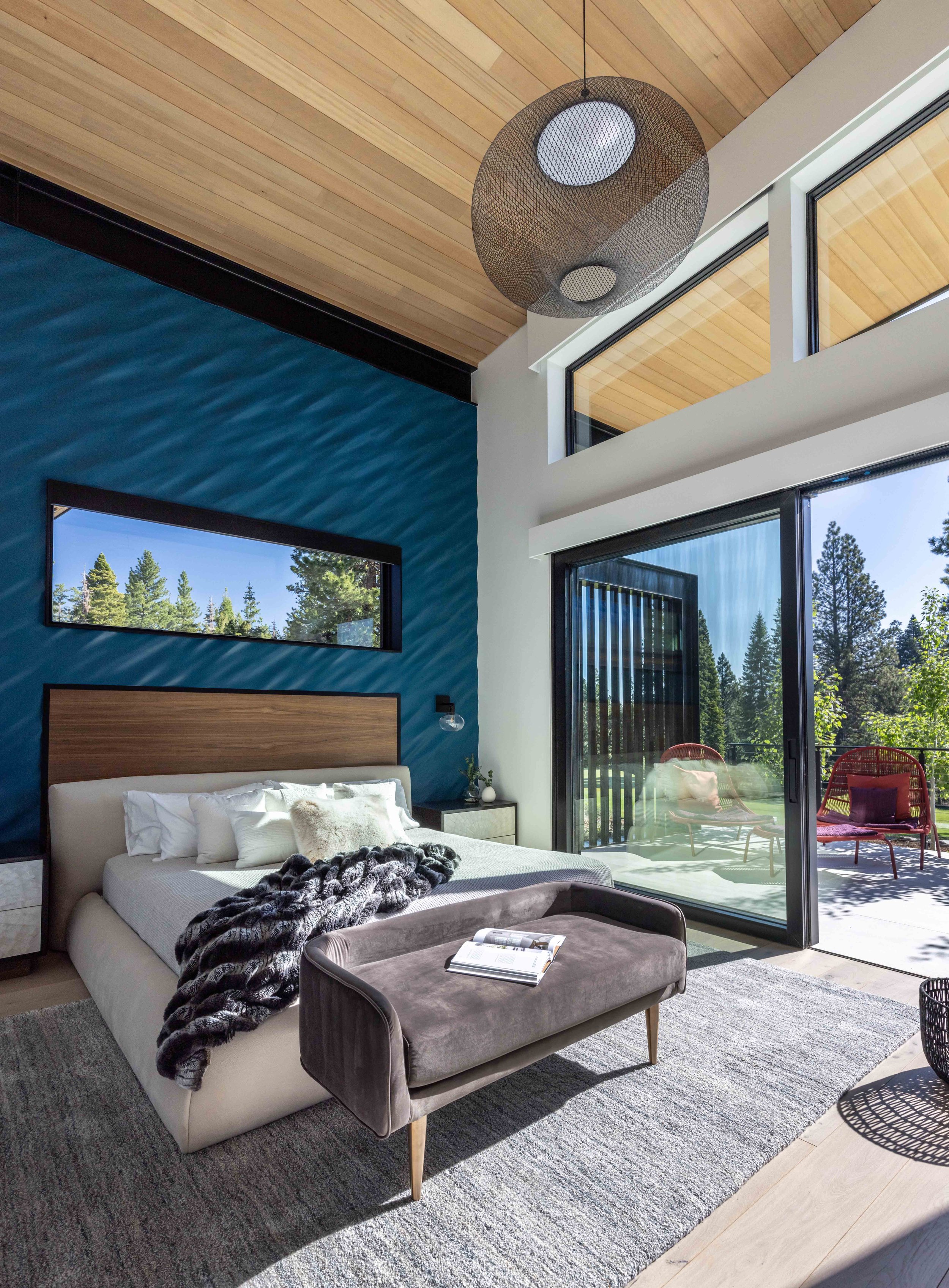 Modern bedroom with blue accent wall, large sliding glass door opening to balcony, wooden ceiling, and a bed with white linens and a fur throw.