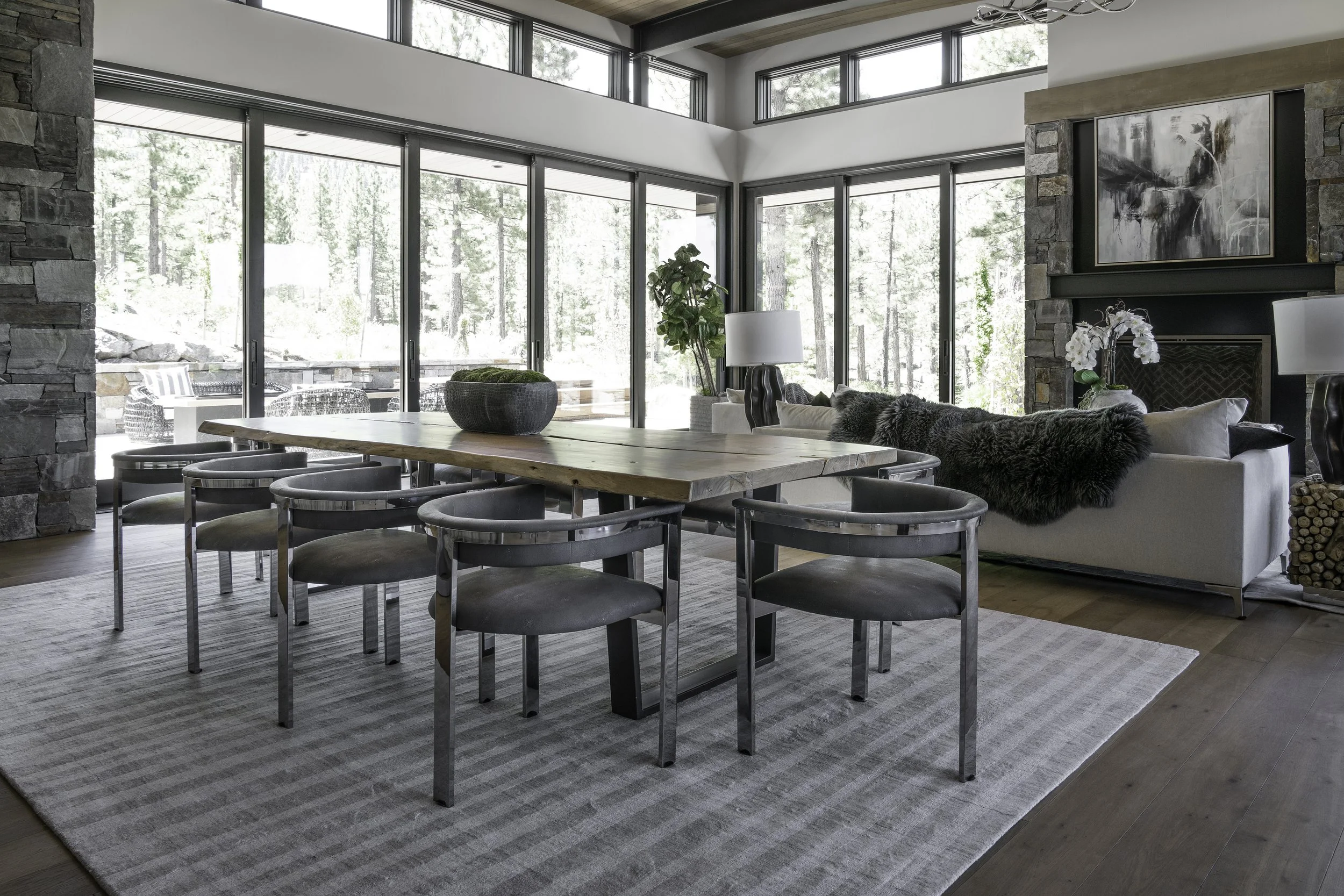 A modern dining and living area with large floor-to-ceiling windows, a rustic wooden dining table with seven chairs, a white sofa with fur and decorative pillows, a stone fireplace with artwork above, and various lamps and plants.