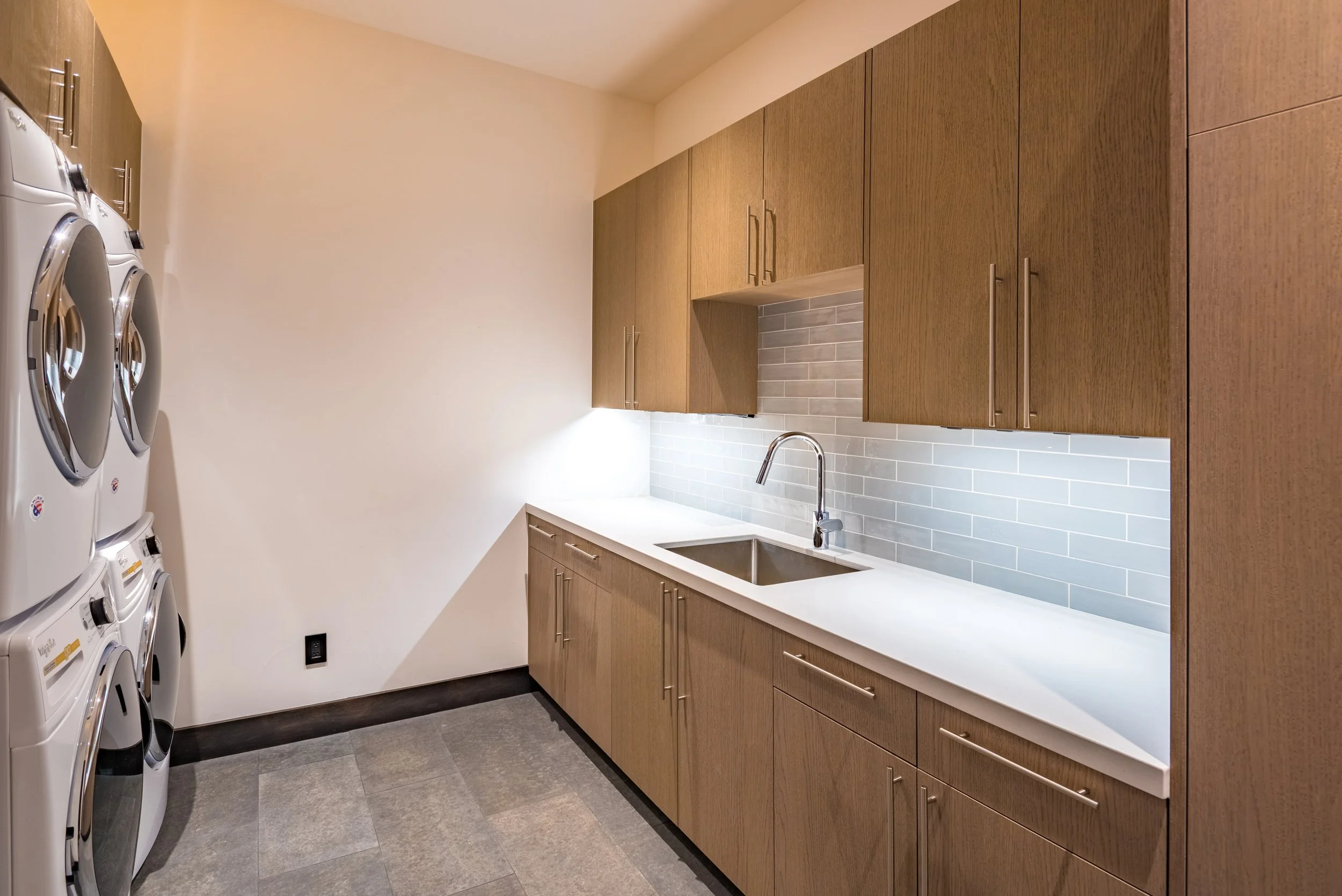 A laundry room with wooden cabinets, a white countertop, a stainless steel sink with a faucet, a gray tile backsplash, and a stack of washers and dryers on the left side.