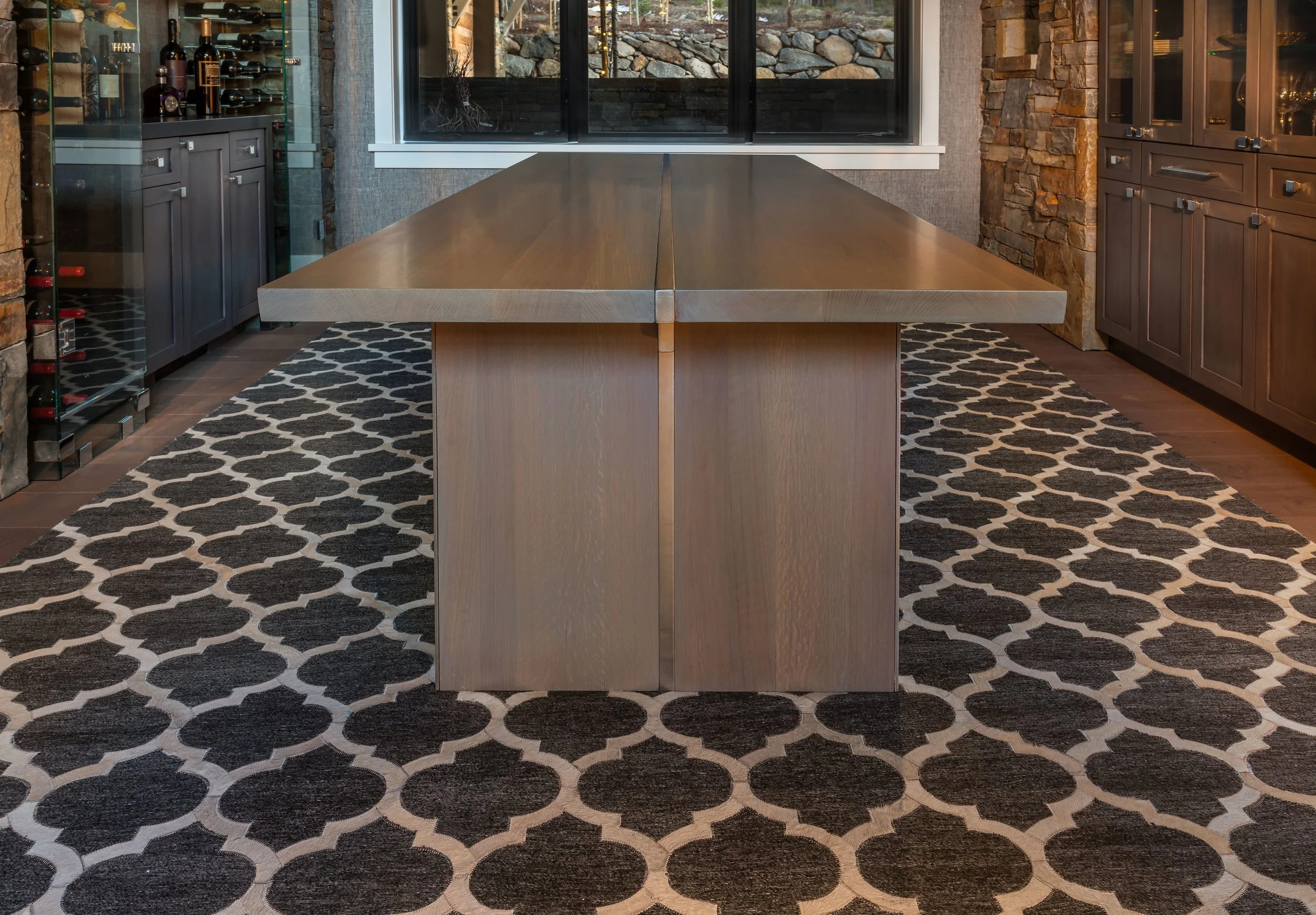 A large wooden dining table in a room with stone walls, a window, and patterned black and white rug.