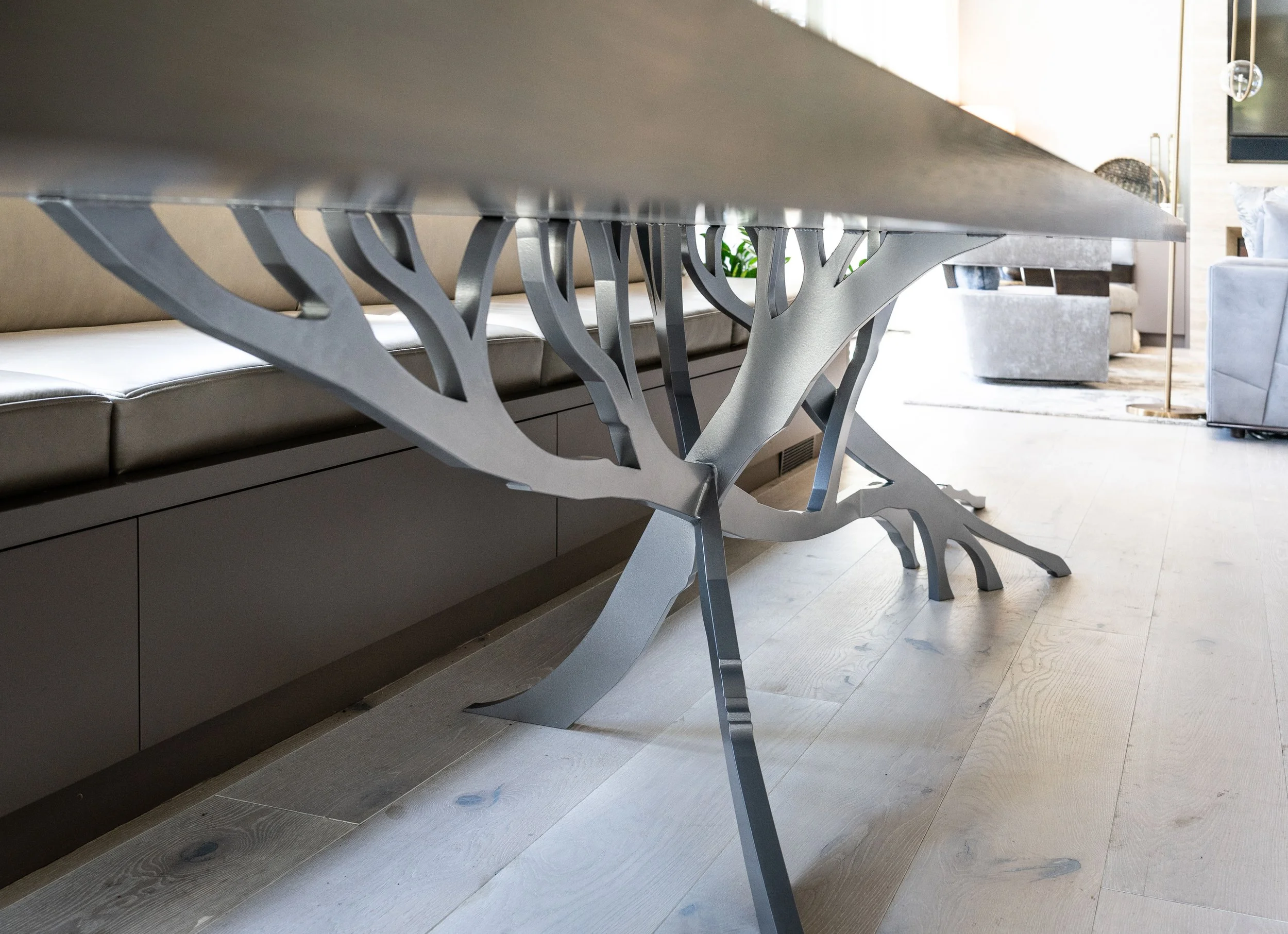 Close-up of a modern dining room table with a unique metallic tree-branch style base, situated in a spacious, well-lit living area with light wood flooring and contemporary furniture.