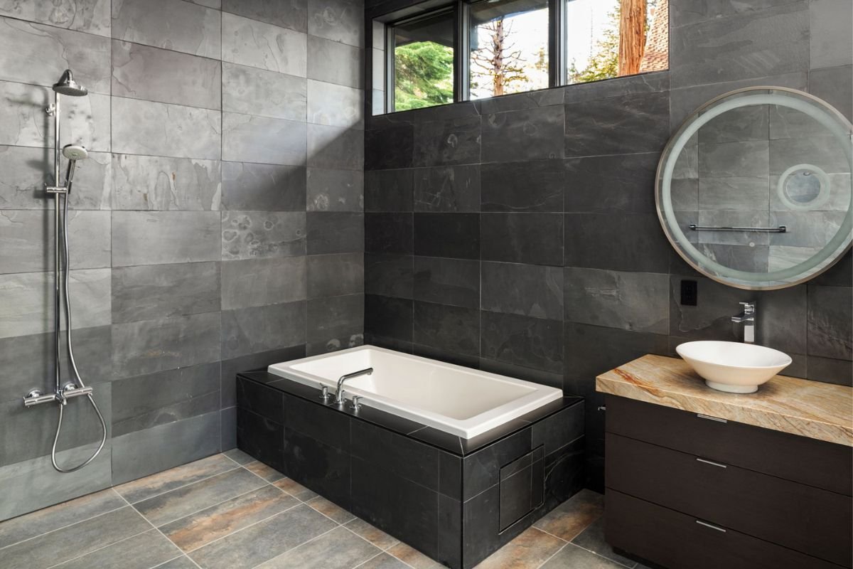 Modern bathroom with a built-in bathtub, a wall-mounted showerhead, a small vanity with a vessel sink, and a large round mirror. The walls and floor are tiled in shades of gray, and there is a window near the ceiling providing natural light.