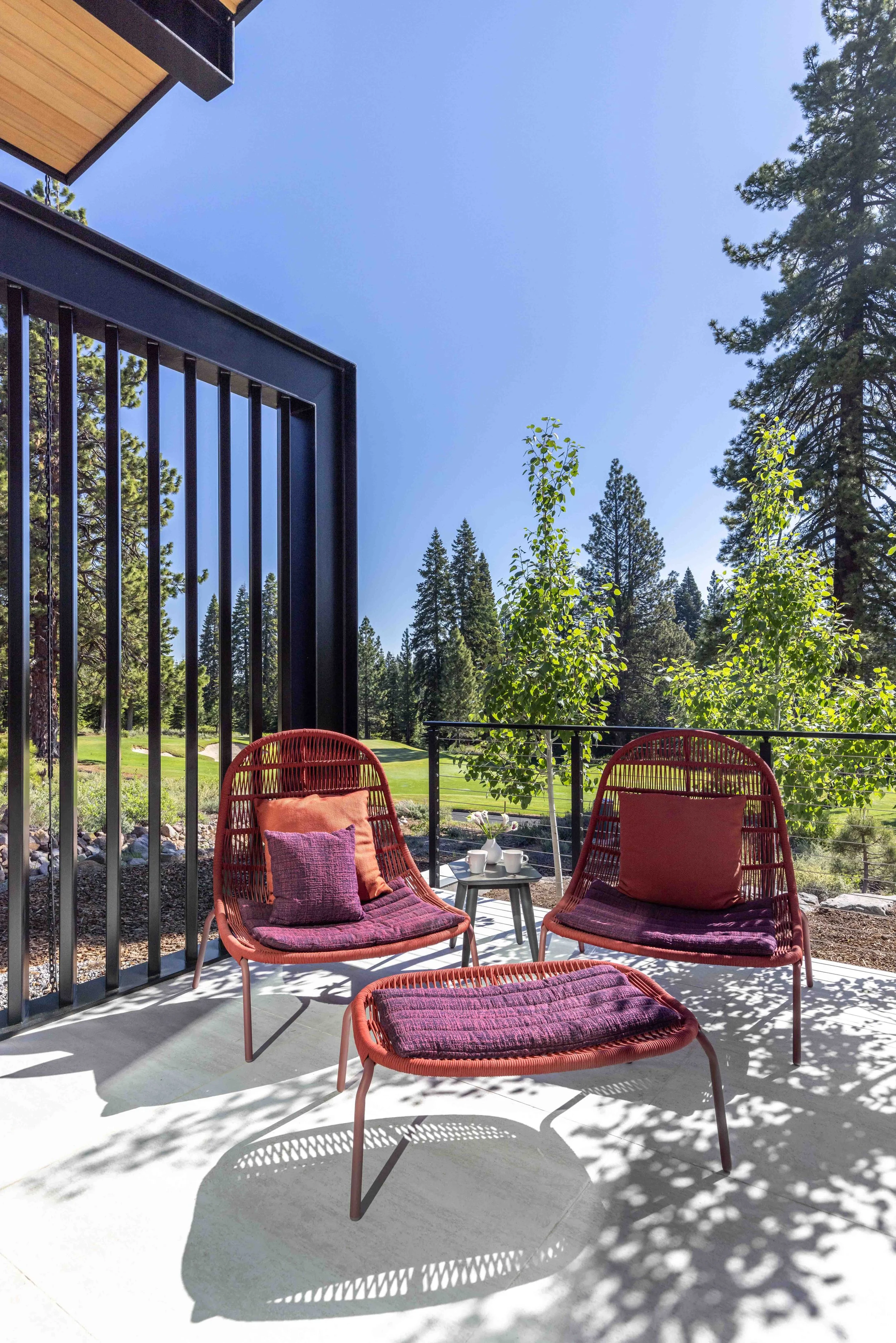 Outdoor patio with two red chairs with purple cushions and a matching footrest, a small round table with cups and flowers, surrounded by trees and green landscape under a clear blue sky.