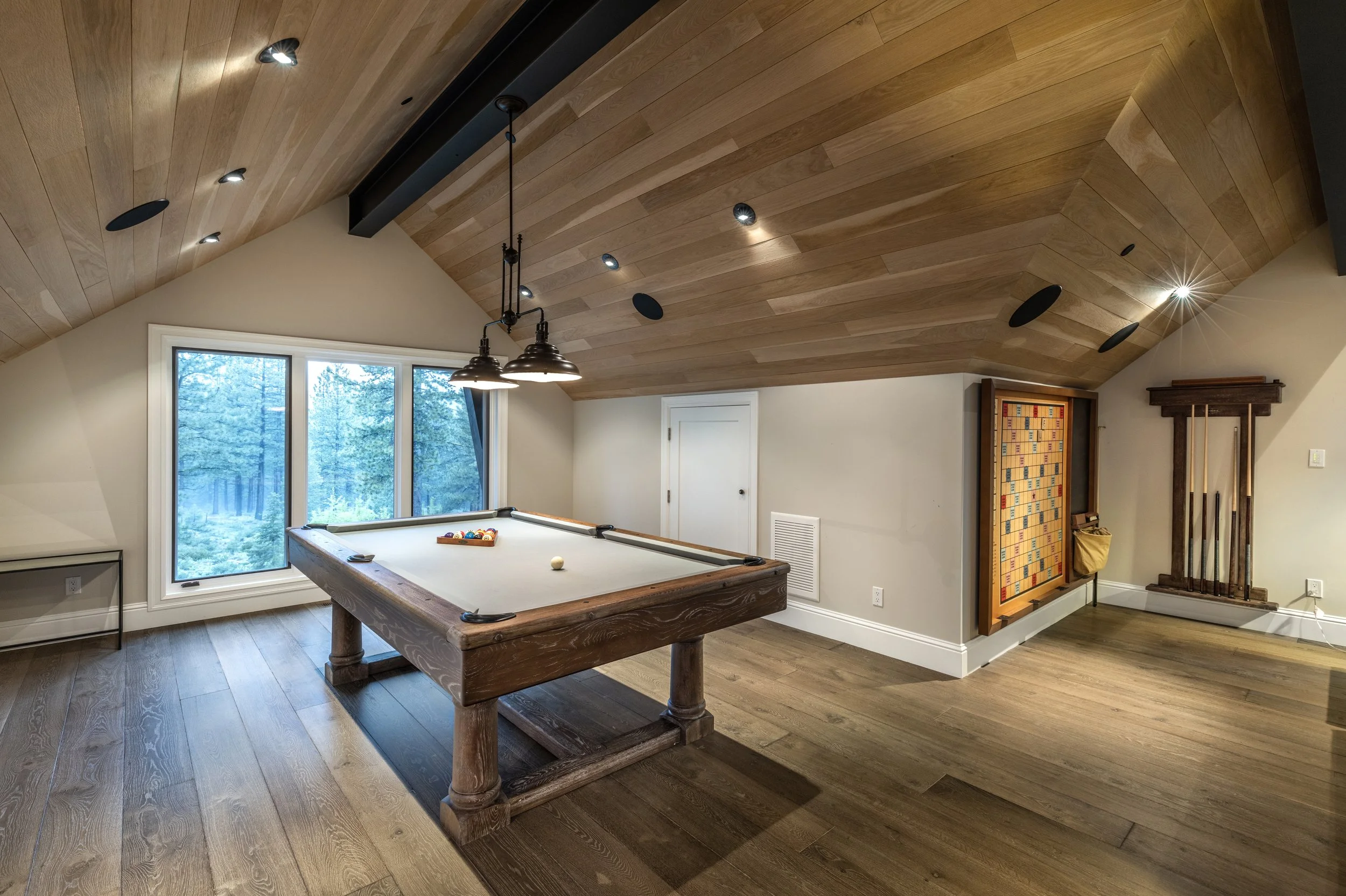 A cozy game room with wooden ceilings and floors, featuring a pool table in the center near a large window with a forest view, and a cue rack on the wall.