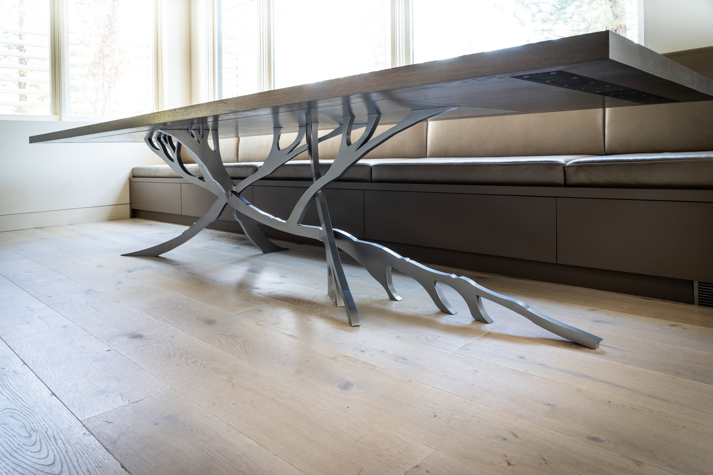 A modern dining table with a wooden top and sculptural metal legs in a bright room with large windows and light-colored hardwood floors.