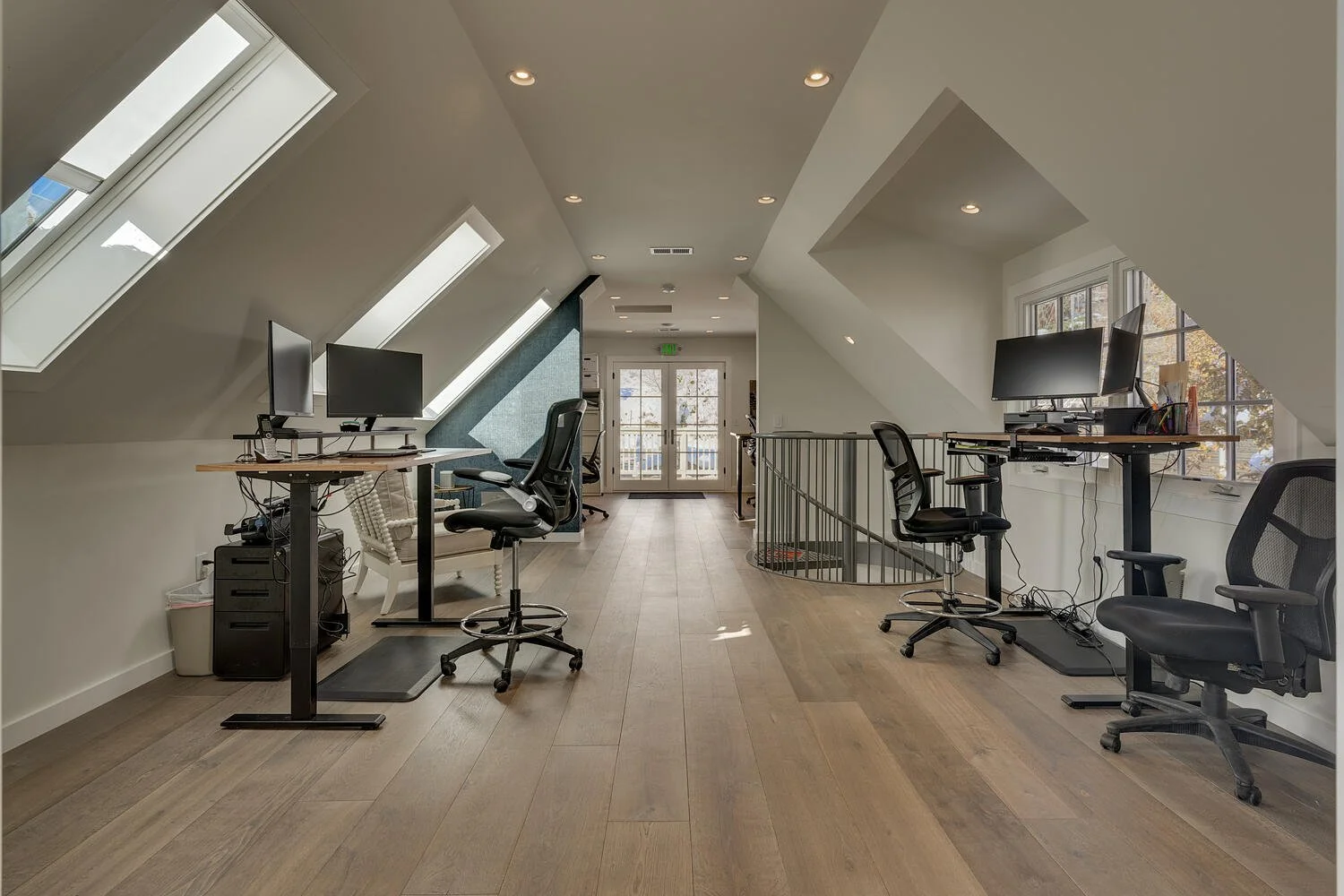 Home office space with four black office chairs and multiple computer monitors, under sloped ceiling with skylights, and a door at the back leading outside.
