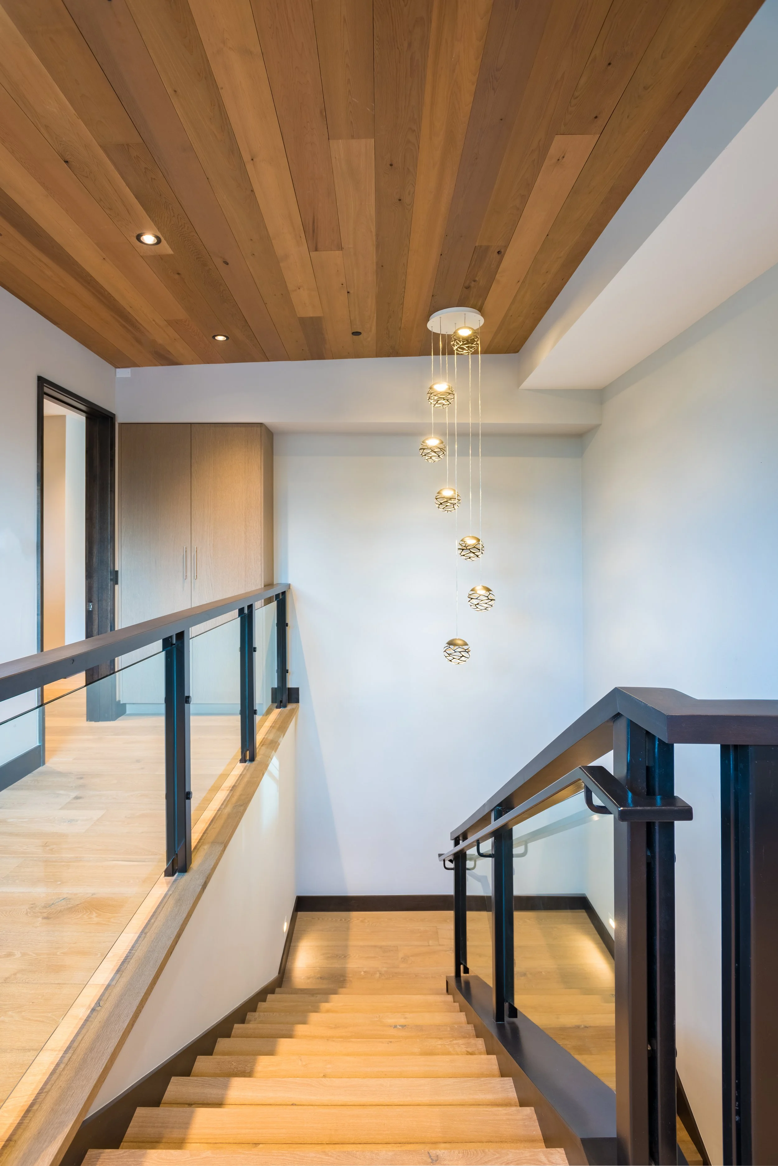 Interior view of a staircase landing with wooden stairs, a wooden ceiling, modern hanging pendant lights, a black railing, and a wooden cabinet.