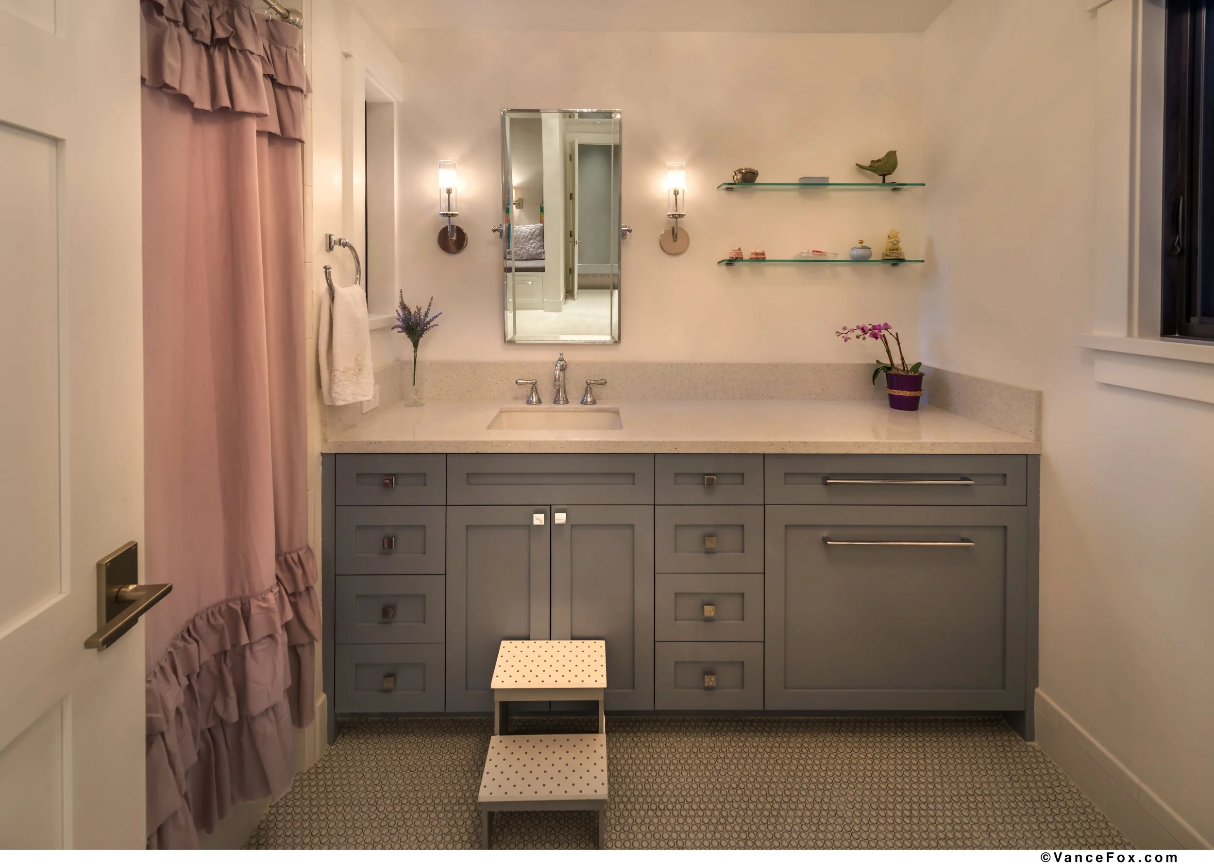 A bathroom with a gray vanity, a white countertop, a rectangular mirror, two wall lights, a window, pink curtains, and decorative shelves with small ornaments. There is a step stool in front of the vanity and a towel hanging on a ring.