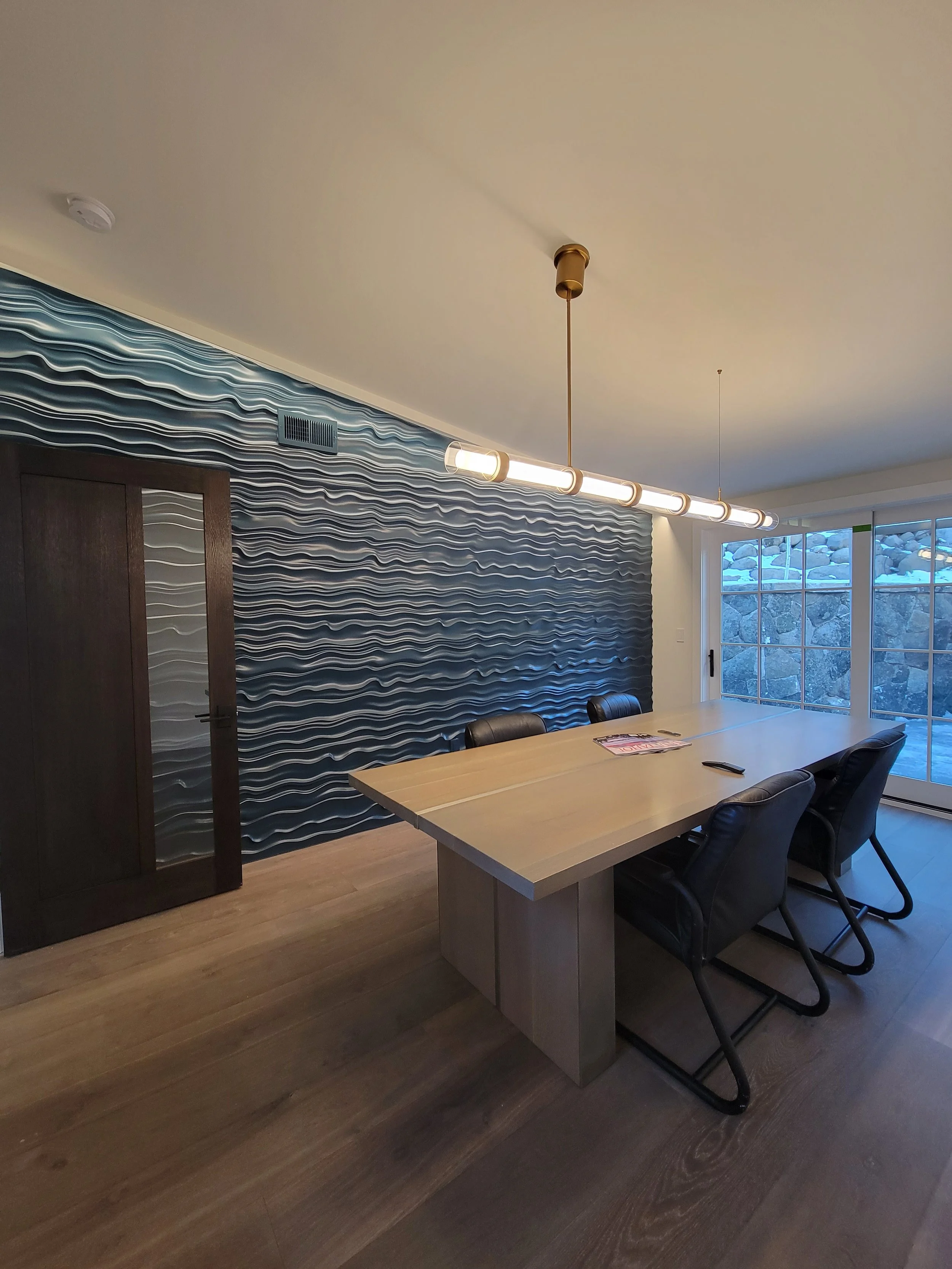 Dining room with a wooden table, black chairs, a decorative wavy blue and white textured wall, a large window with sliding glass door, and a modern linear chandelier hanging from the ceiling.