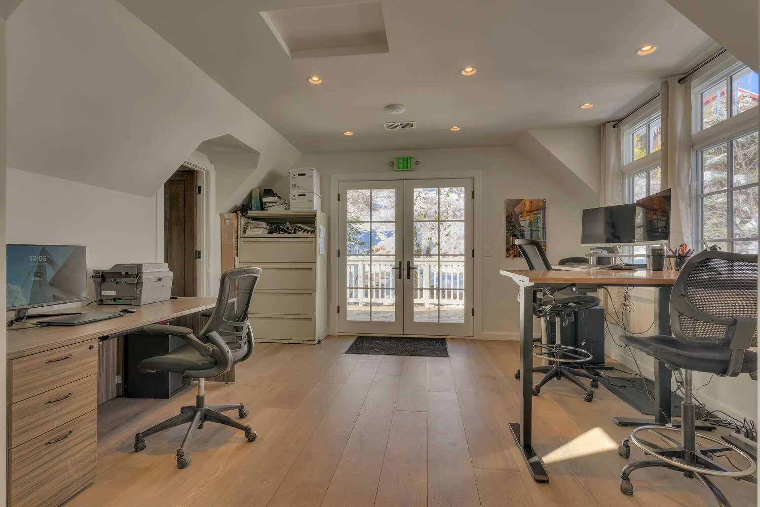 Office room with two desks, each with computers and office chairs, large windows, and a view of a deck outside, with natural sunlight and light wood floors.