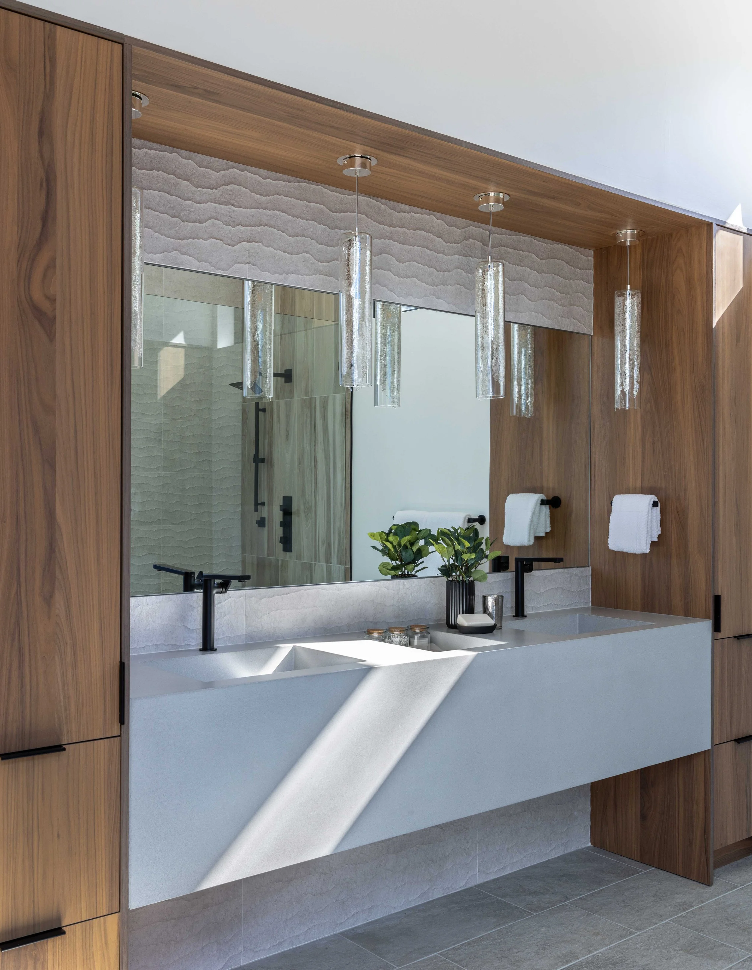 Modern bathroom with double white countertop sink, black faucets, wooden cabinetry, large mirror, pendant lights, and a shower in the background.