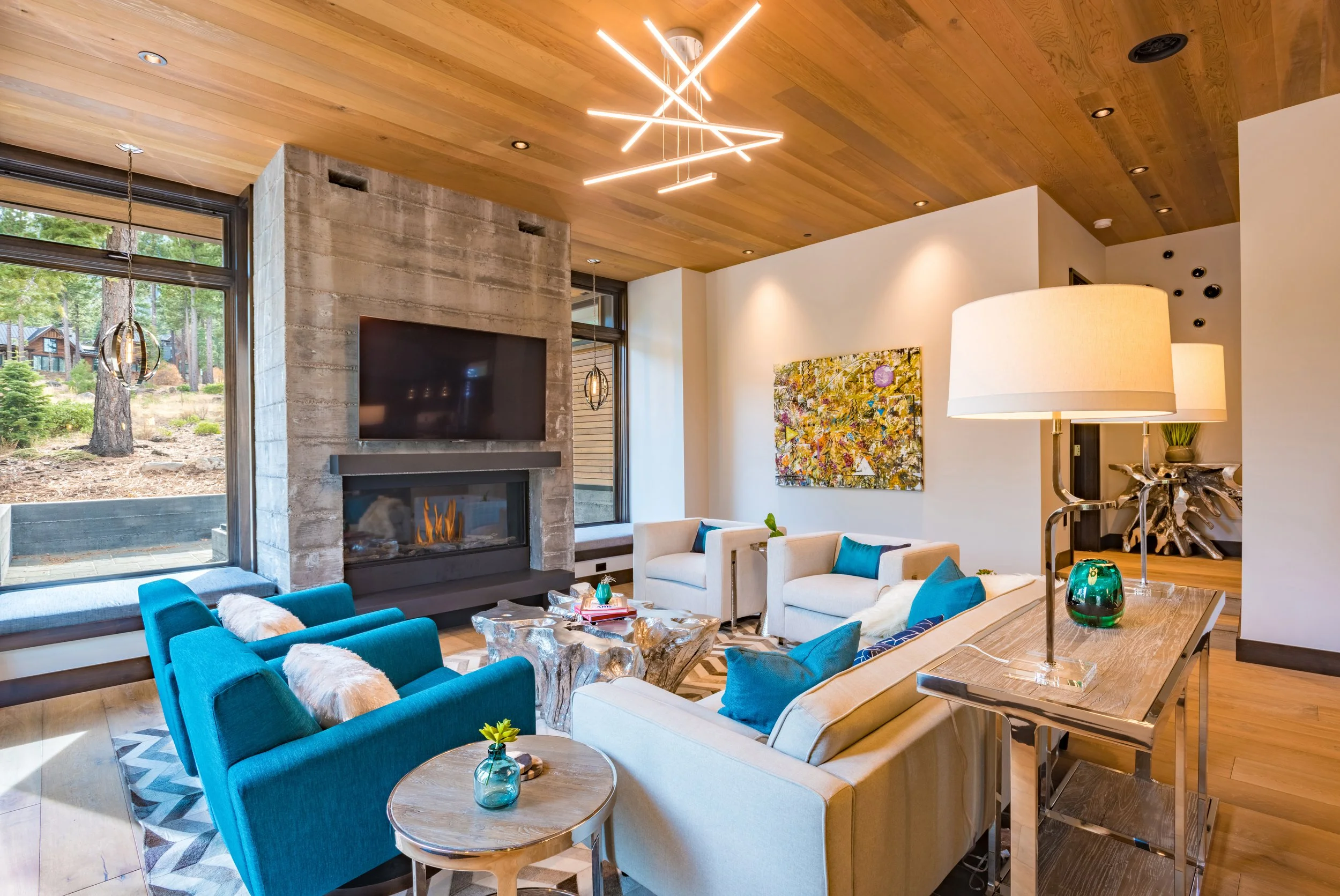 Modern living room with a stone fireplace, blue and white seating, wall art, and large windows showing an outdoor landscape.