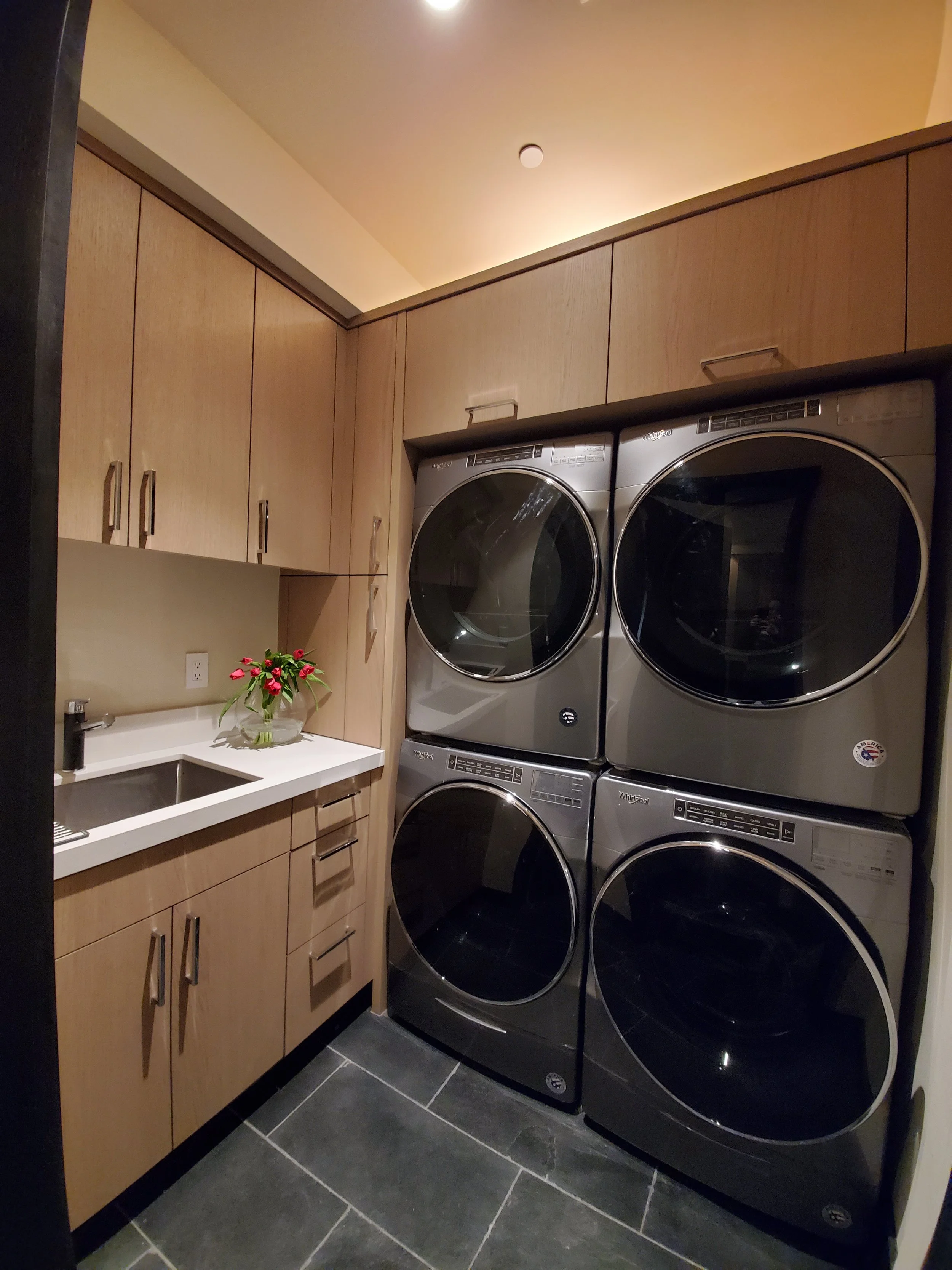 A laundry room with a stack of four silver front-loading washing machines and dryers, wooden cabinets, a small white sink with a black faucet, and a vase with pink and red flowers.