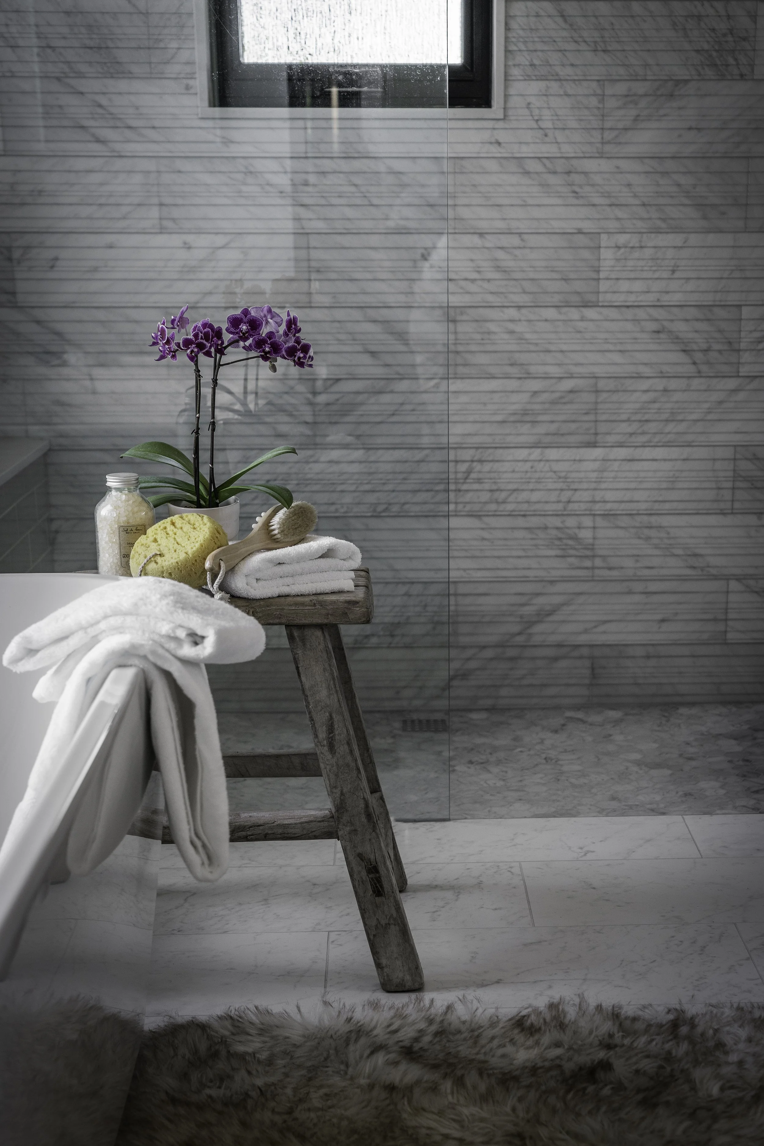 A bathroom with a wooden stool holding folded white towels, a purple orchid plant, a bath brush, a sponge, bath salts, and a bottle of bath products; a fluffy rug on the floor and a marble shower wall with a small window.