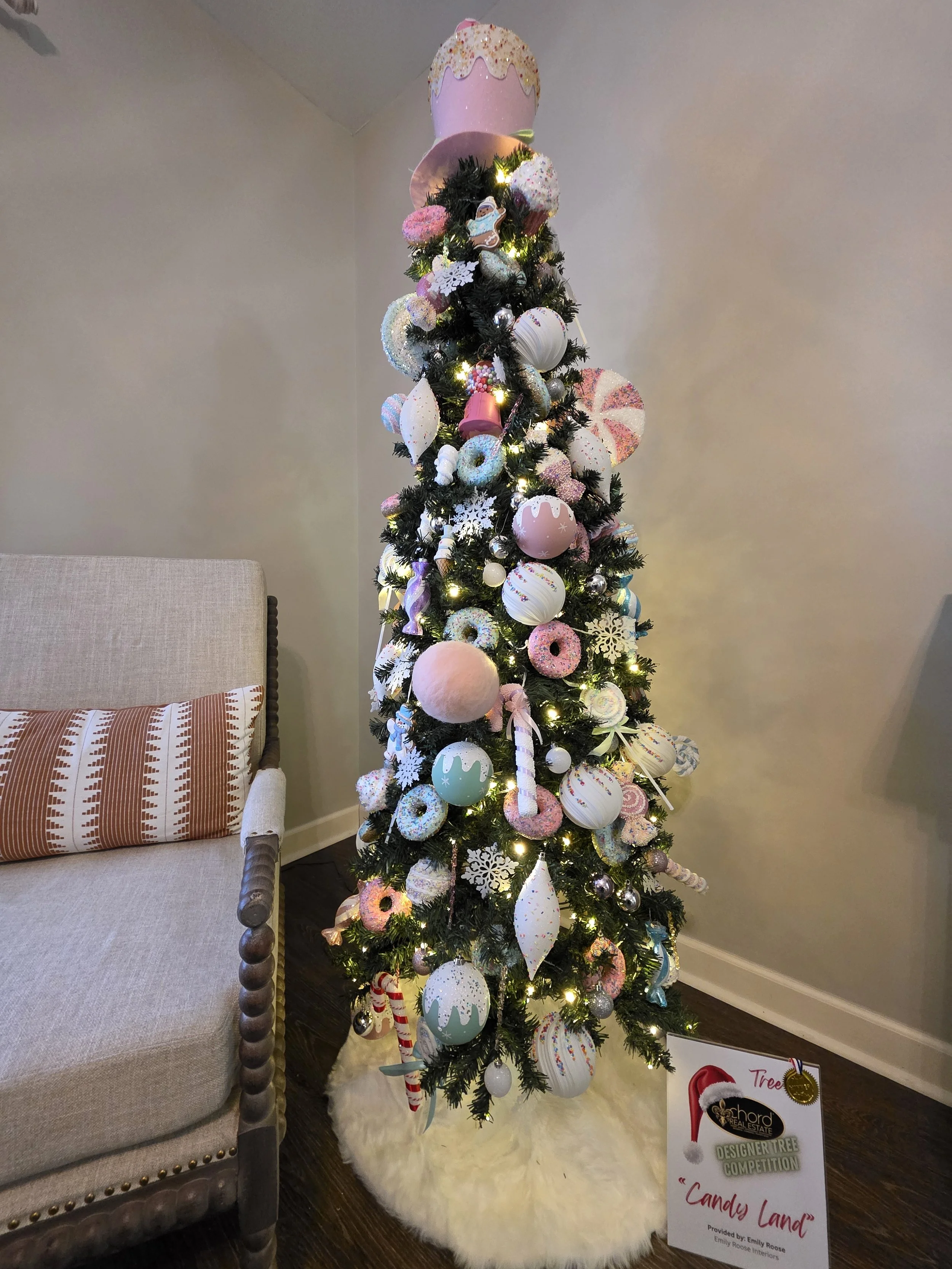 Decorated Christmas tree with pink, white, and pastel ornaments and lights, topped with a pink hat, standing on a white furry tree skirt next to a beige sofa