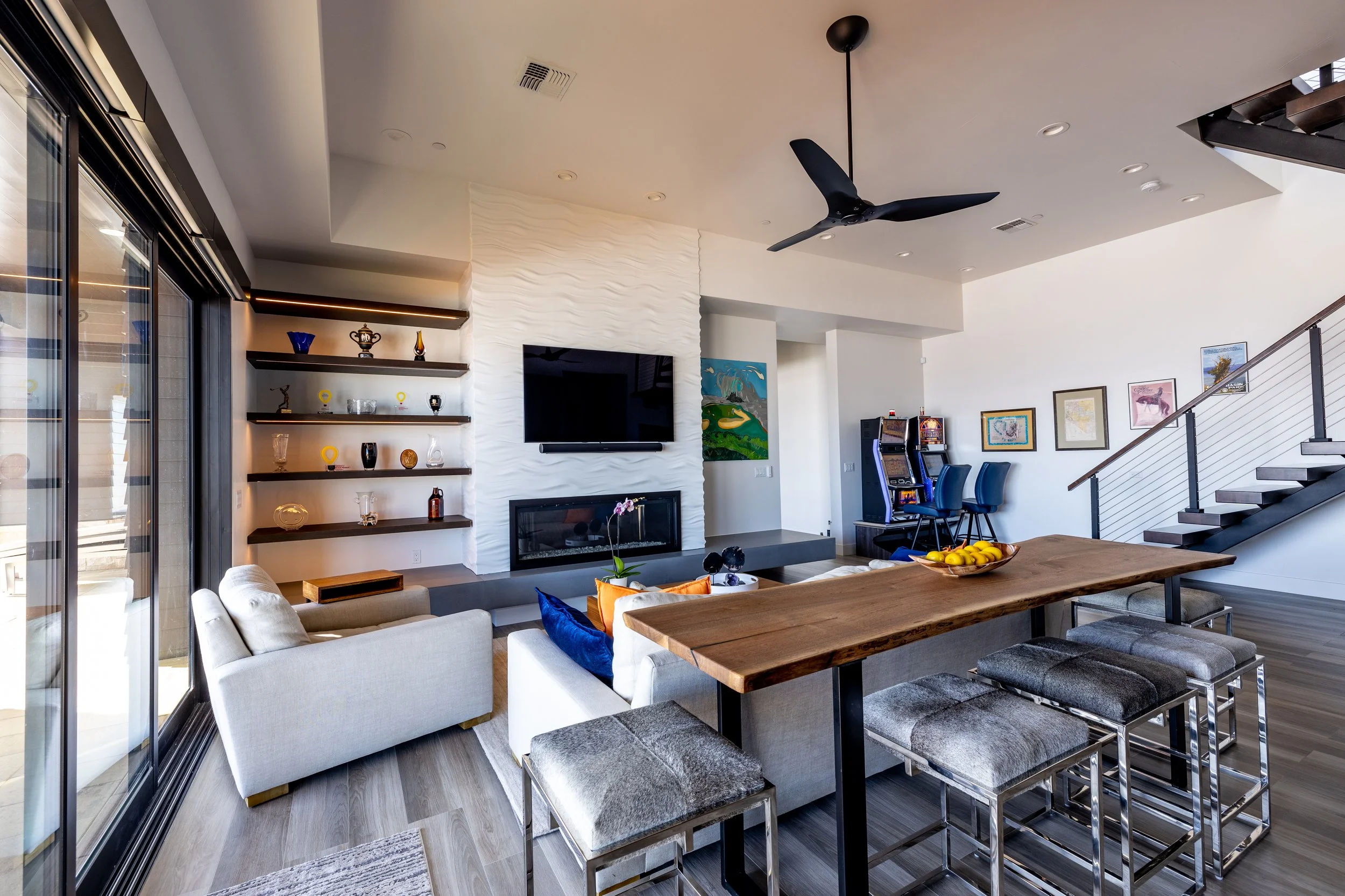 Interior of a modern living room with a white sectional sofa, a wooden dining table with stools, a white textured wall with a mounted TV and a fireplace, shelves with decorative items, a staircase, and a corner with arcade game machines.