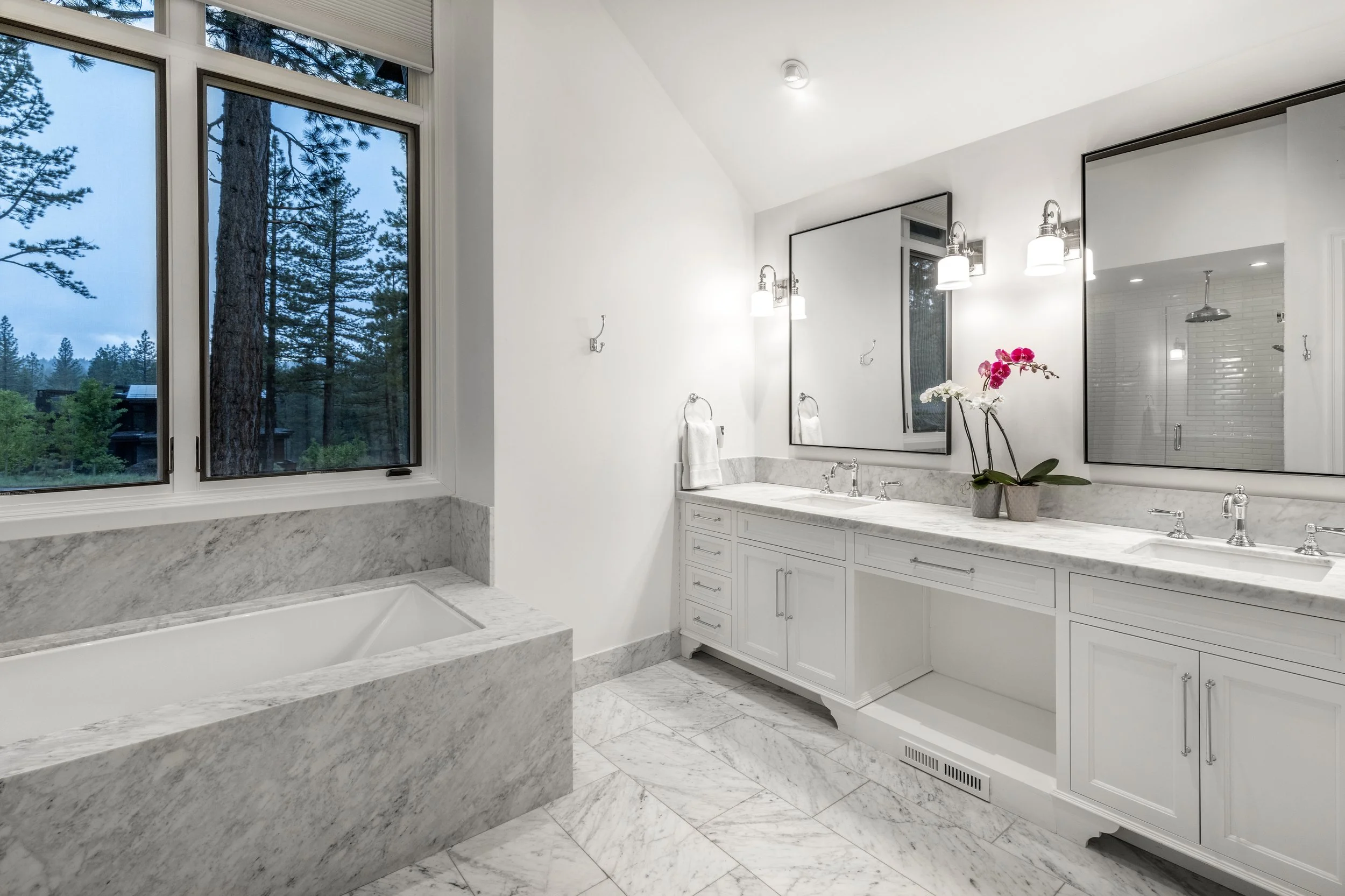 Modern bathroom with double sink vanity, large mirror, and a window overlooking trees. Features white cabinetry, marble countertops, and a built-in bathtub with marble surround.