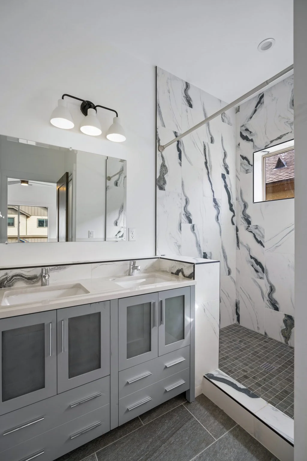 Modern bathroom with double vanity, large mirror, three-light fixture, marble wall tiles, small window, and walk-in shower with gray tile floor.