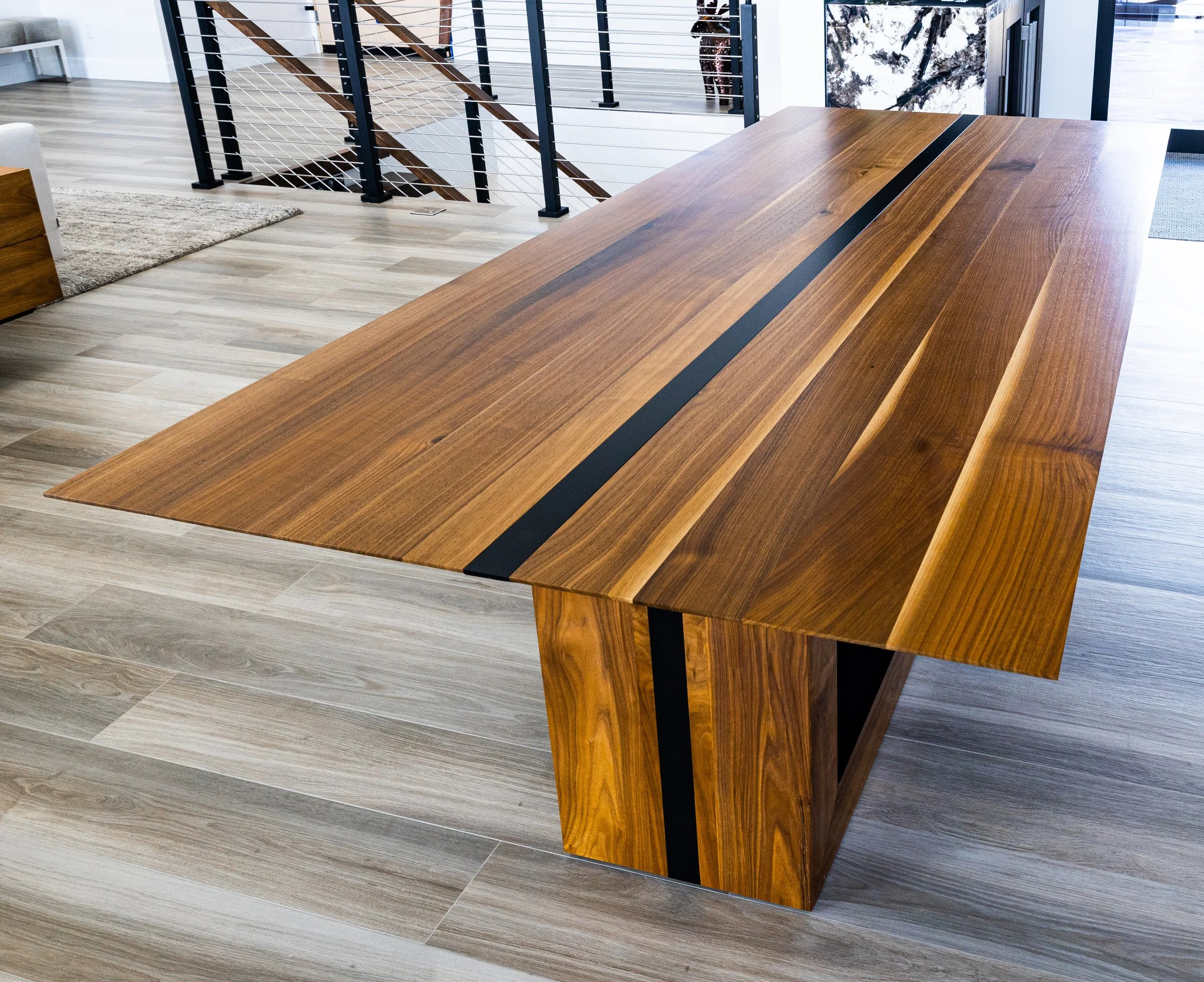A large wooden dining table with black accents in a modern room with wood flooring and staircase.