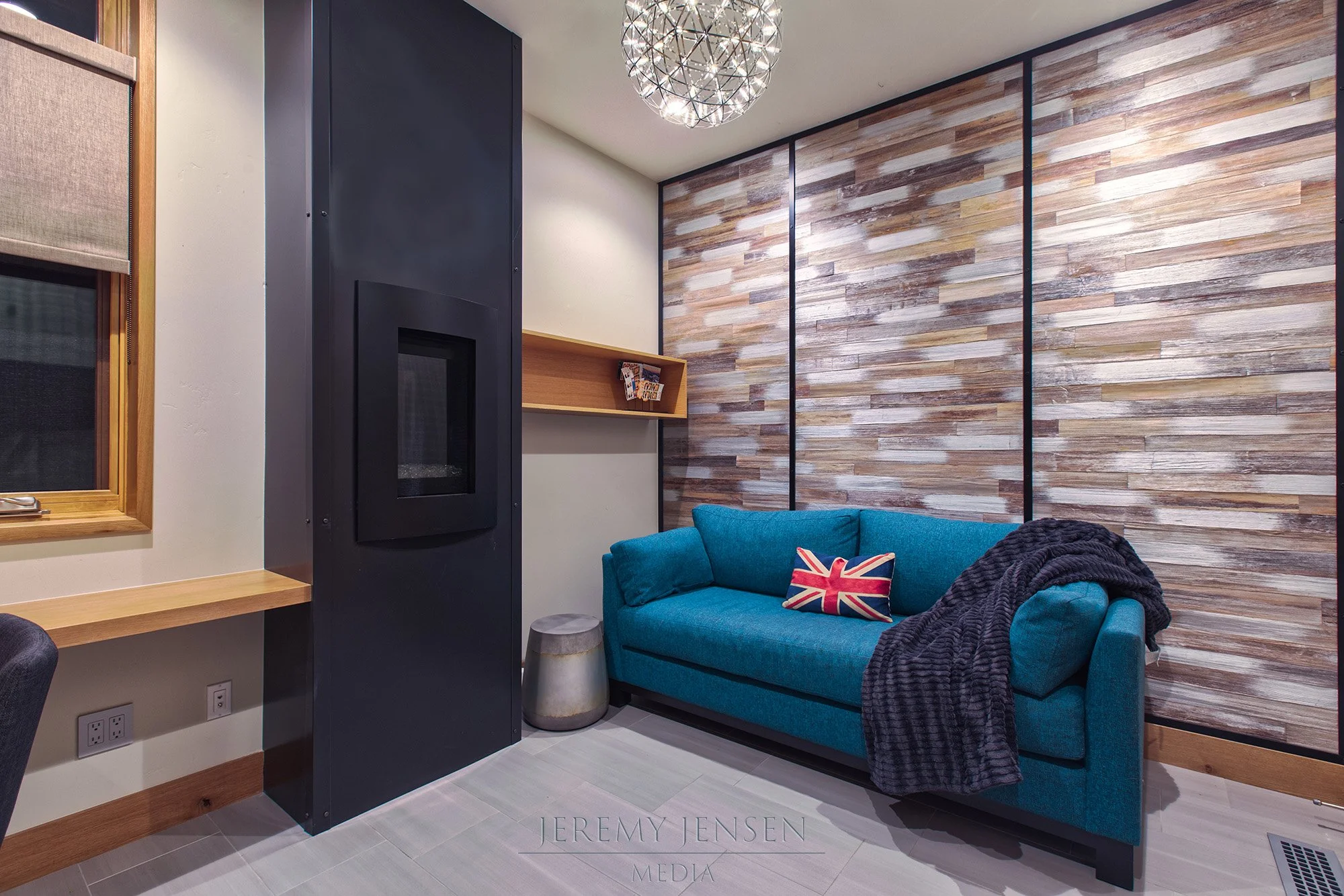 A small cozy living room with a bright blue couch, a Union Jack pillow, a gray throw blanket, a wooden window frame with a shade, a black fireplace wall, a wooden shelf, a textured wooden accent wall, a spherical chandelier, and a metallic side table