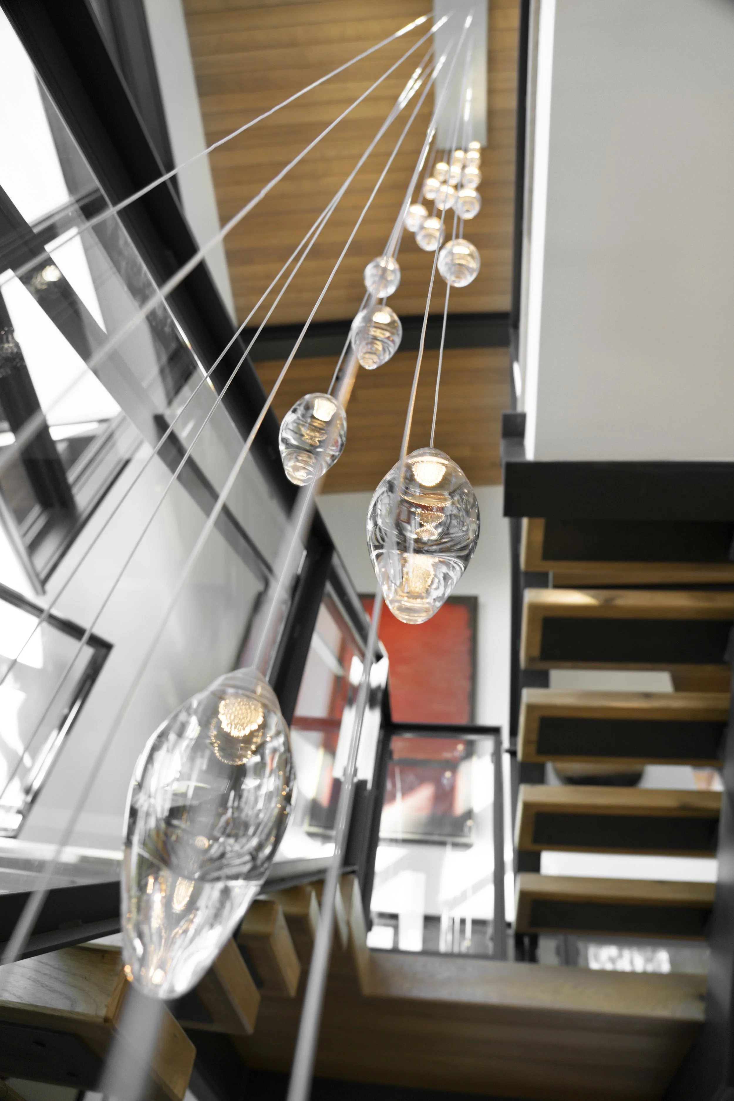 A modern interior staircase with hanging pendant lights made of glass, viewed from below, showing a wooden ceiling and surrounding walls with artwork.