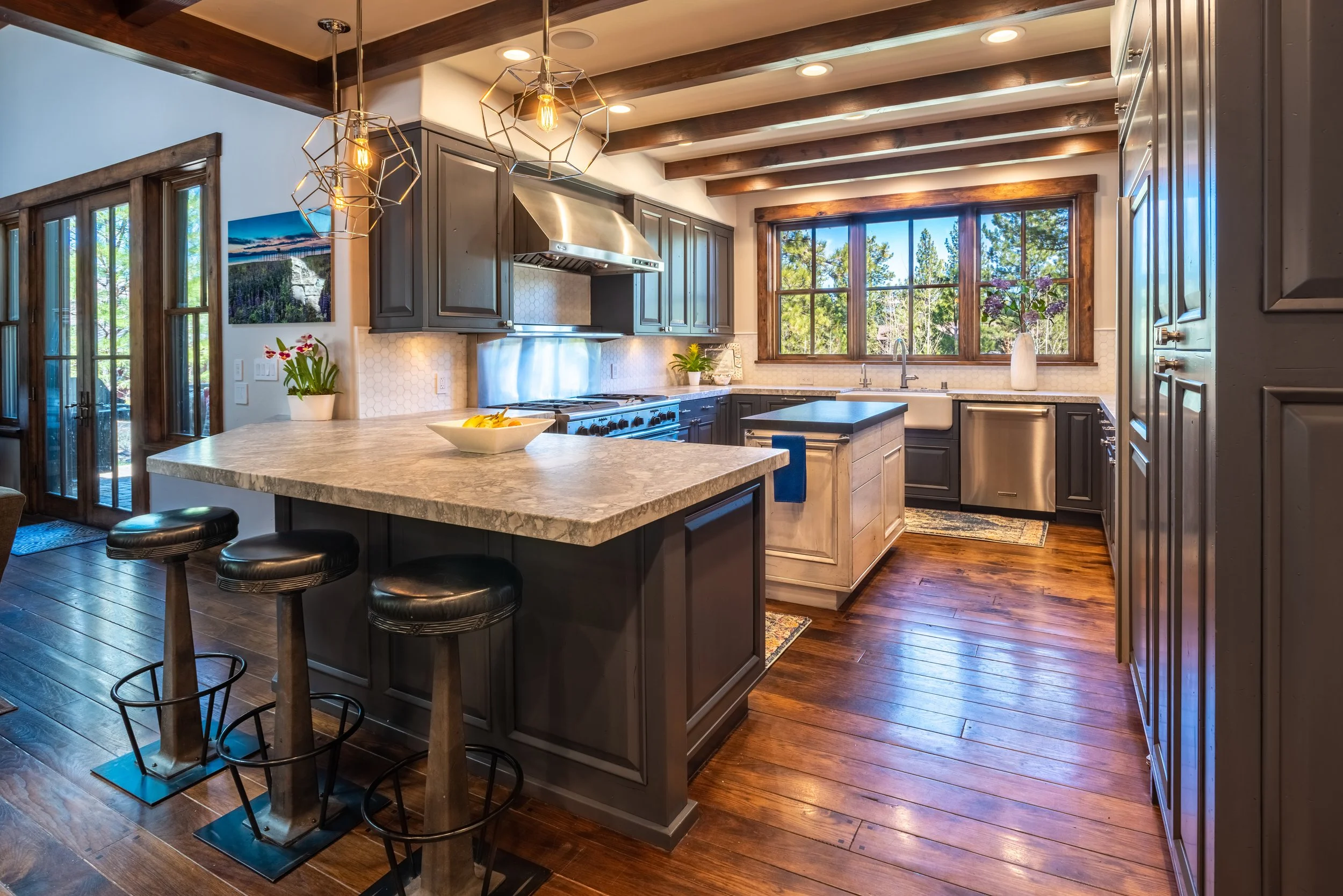 Modern kitchen with gray cabinetry, a large island with a marble countertop, bar stools, and stainless steel appliances. Large window over the sink offers a view of trees outside. Pendant lights hang above the island, and wooden beams decorate the ce