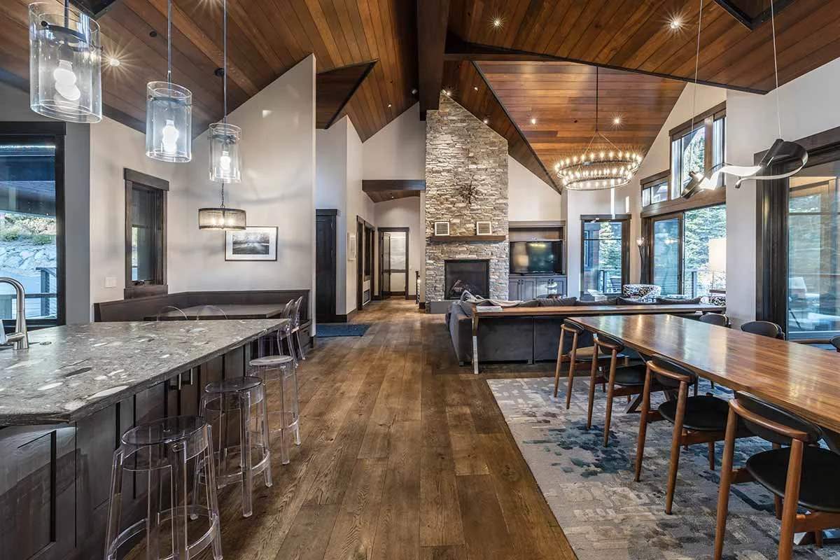 Open-concept living and dining room with wood-paneled vaulted ceiling, large windows, stone fireplace, modern chandelier, and mixed seating areas.