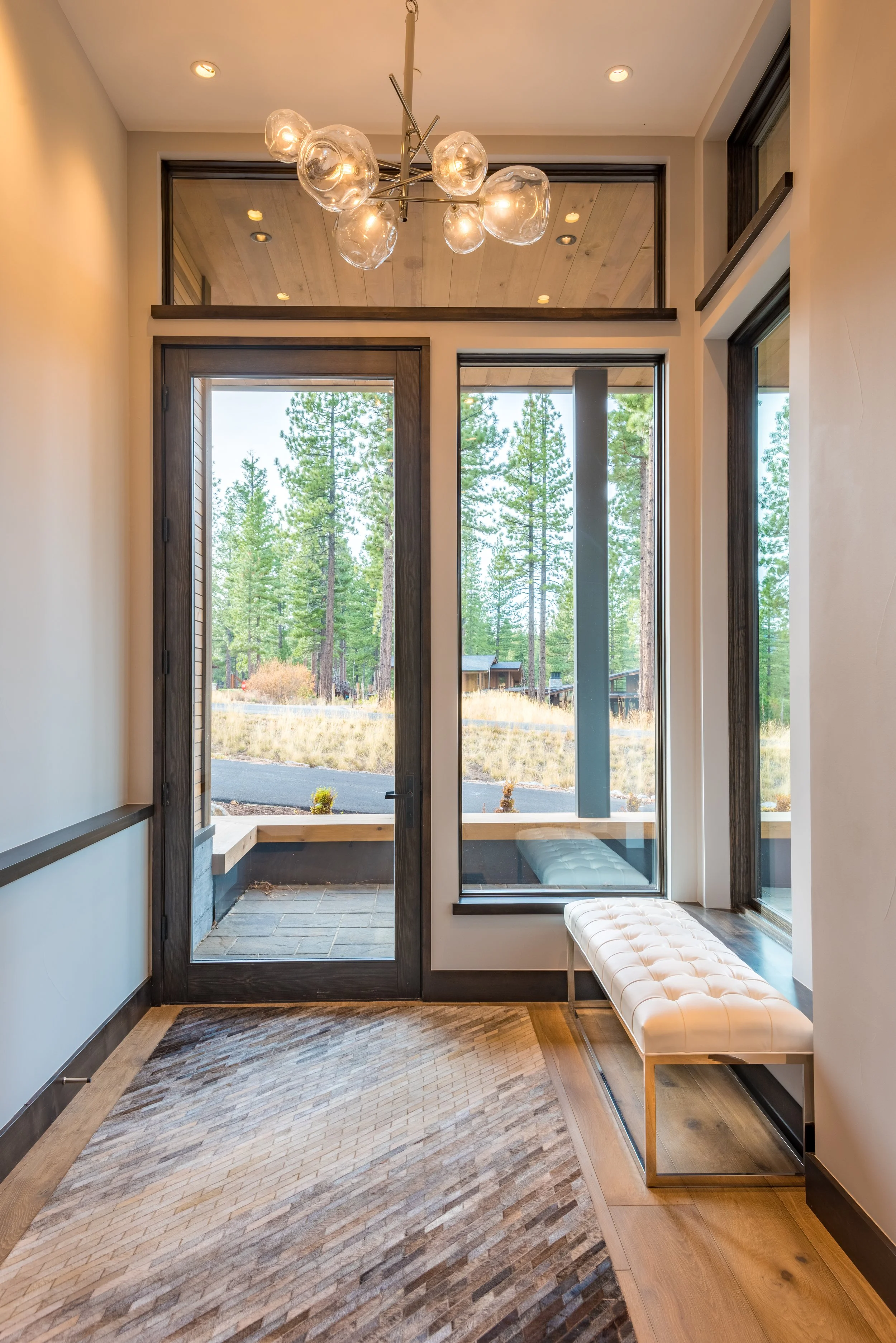 Interior view of a modern entryway with large glass door and windows overlooking a wooded landscape, a white tufted bench, and a contemporary chandelier with glass globes.
