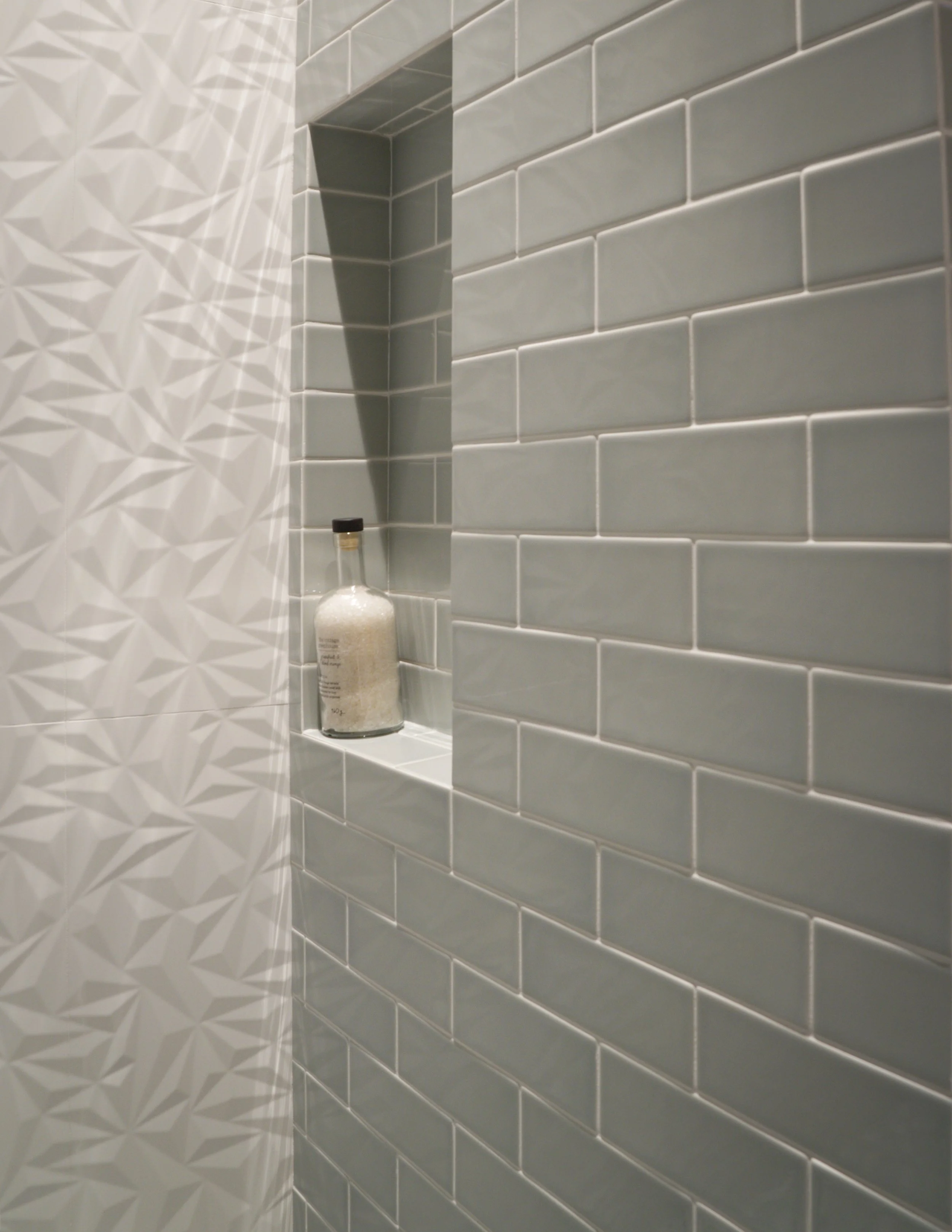 Interior of a shower with gray subway tiles on the walls, a built-in shelf with a bottle of bath salts or soap.