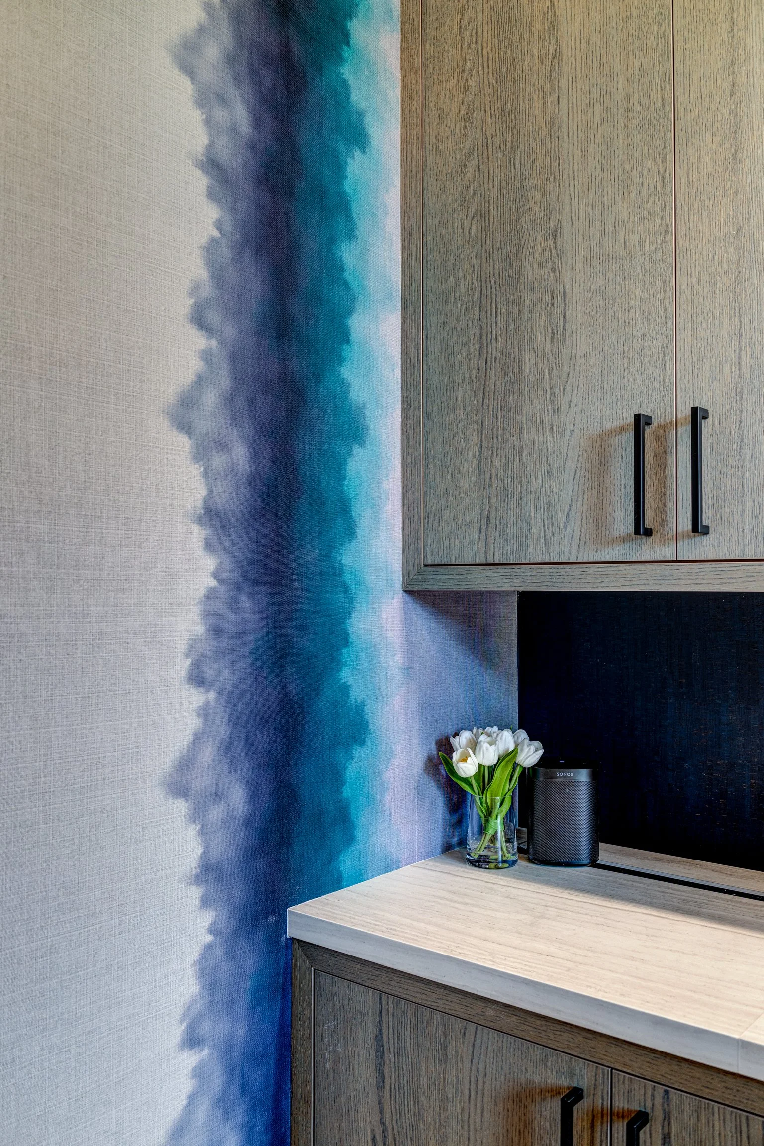 Interior of a kitchen corner with wood cabinets, a countertop, a small bouquet of white tulips in a glass vase, and a black speaker, alongside a wall with a blue watercolor cloud design.